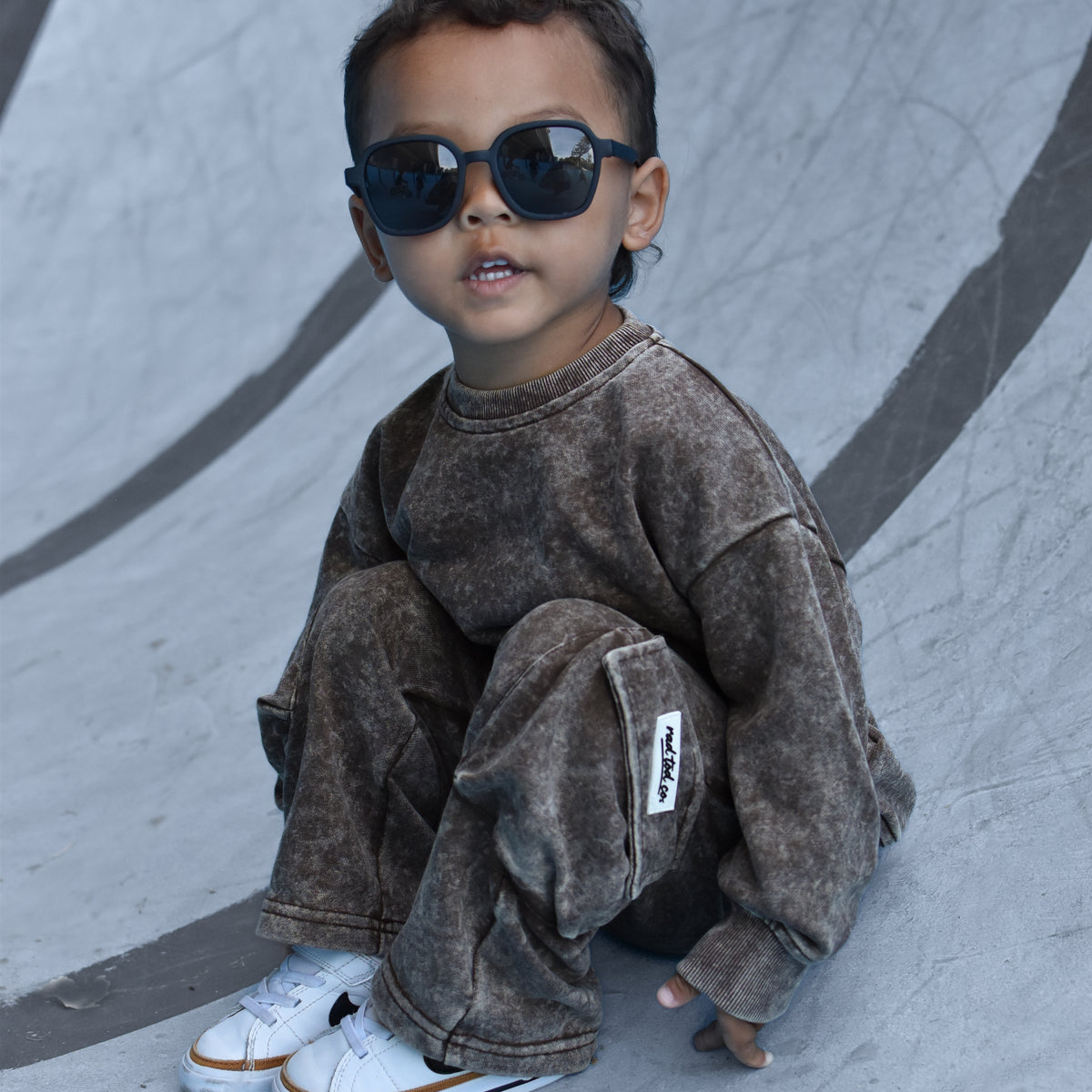 A toddler wearing a matching brown acid-wash sweatshirt and jogger pants set, styled with black sunglasses and white sneakers, sitting on a concrete skatepark ramp.