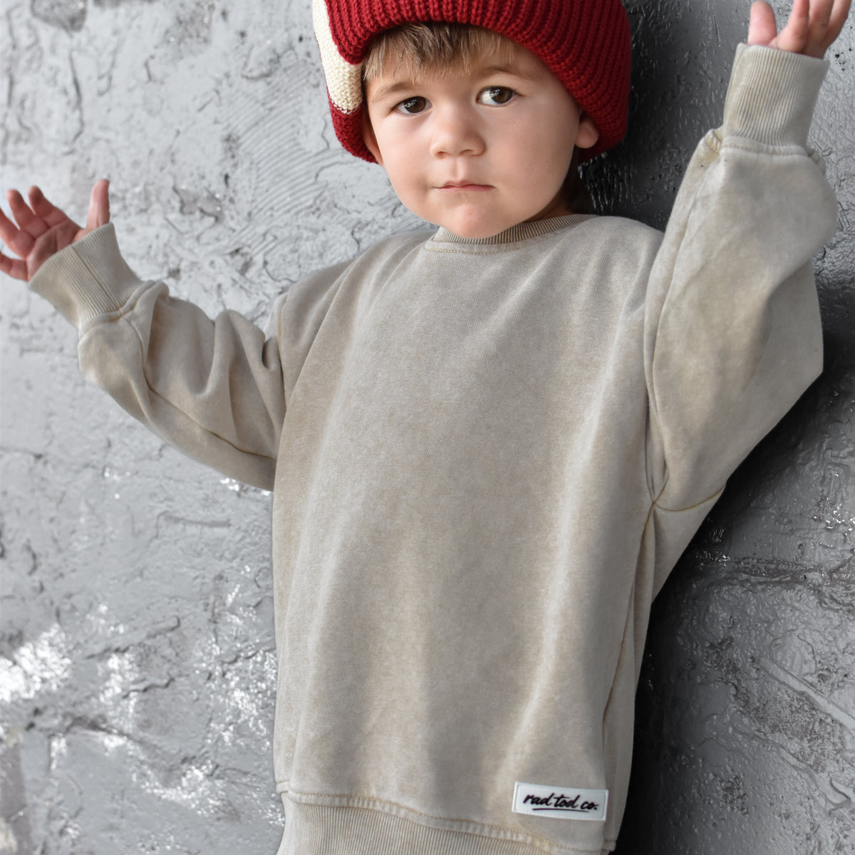 A toddler wearing a beige crewneck sweatshirt and matching pants with a red knit beanie, posing against a textured gray concrete wall.
