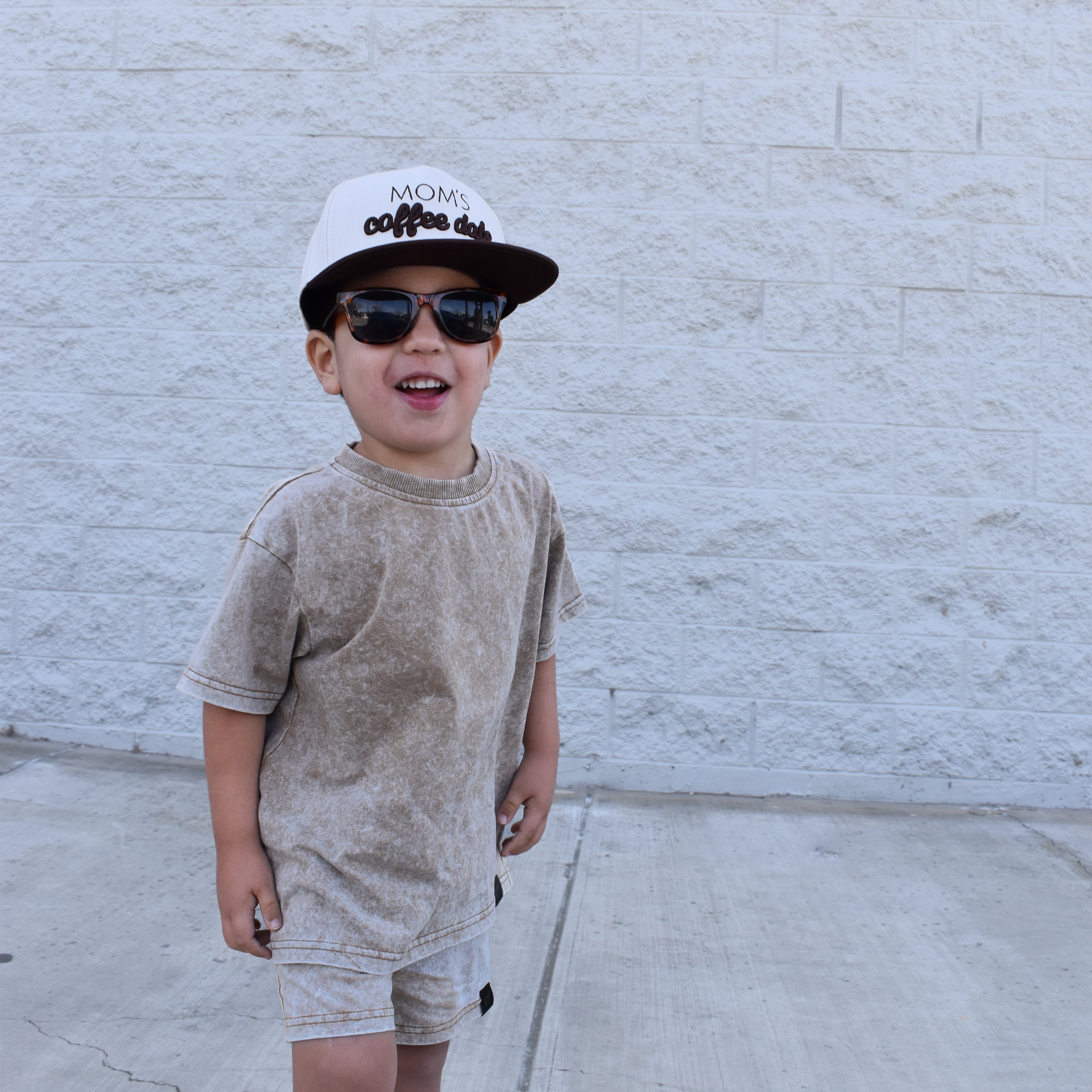 Child wearing sunglasses and a cap against a white brick wall while wearing rad toddlers stone wash sand set