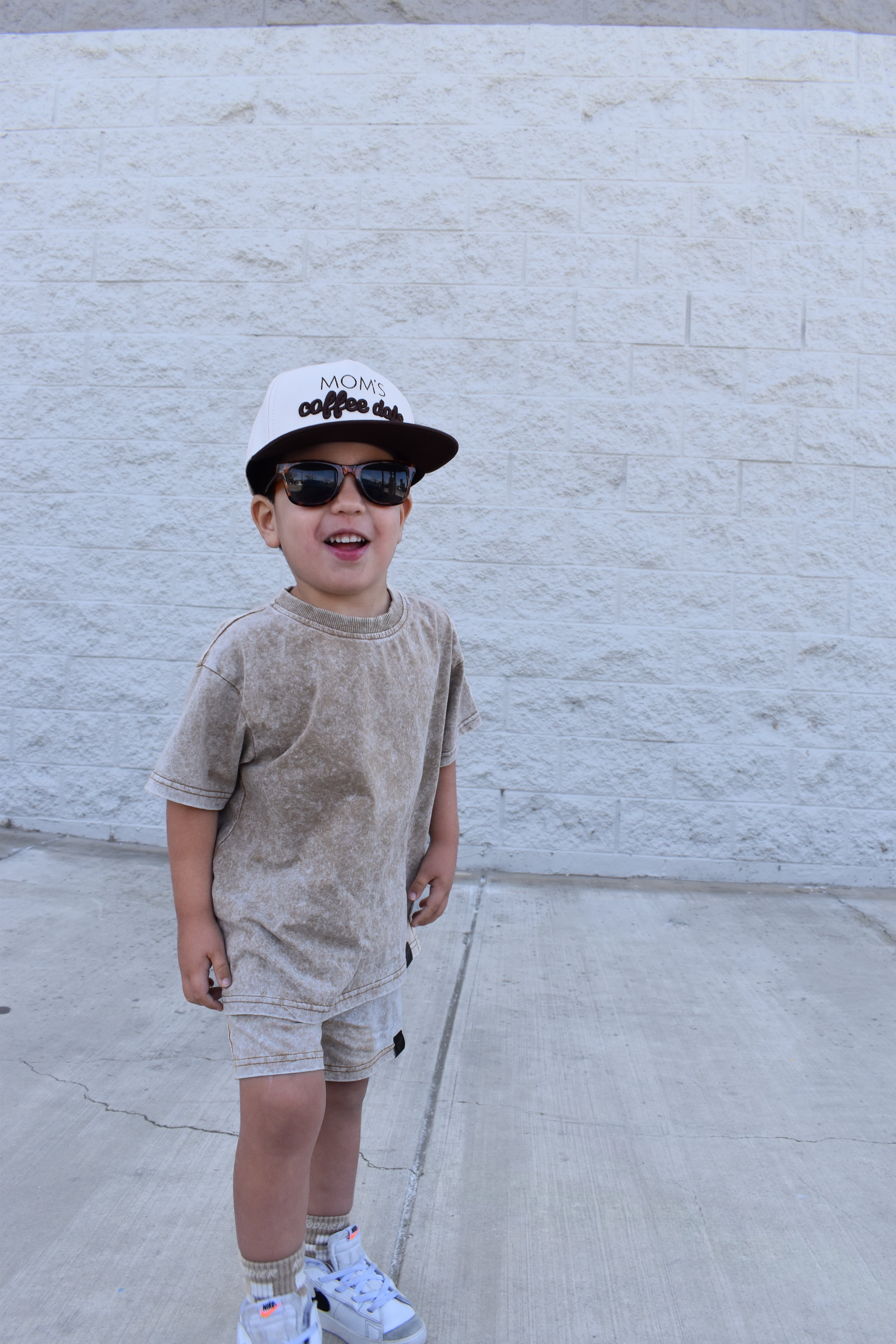 Child wearing sunglasses and a cap against a white brick wall while wearing rad toddlers stone wash sand set