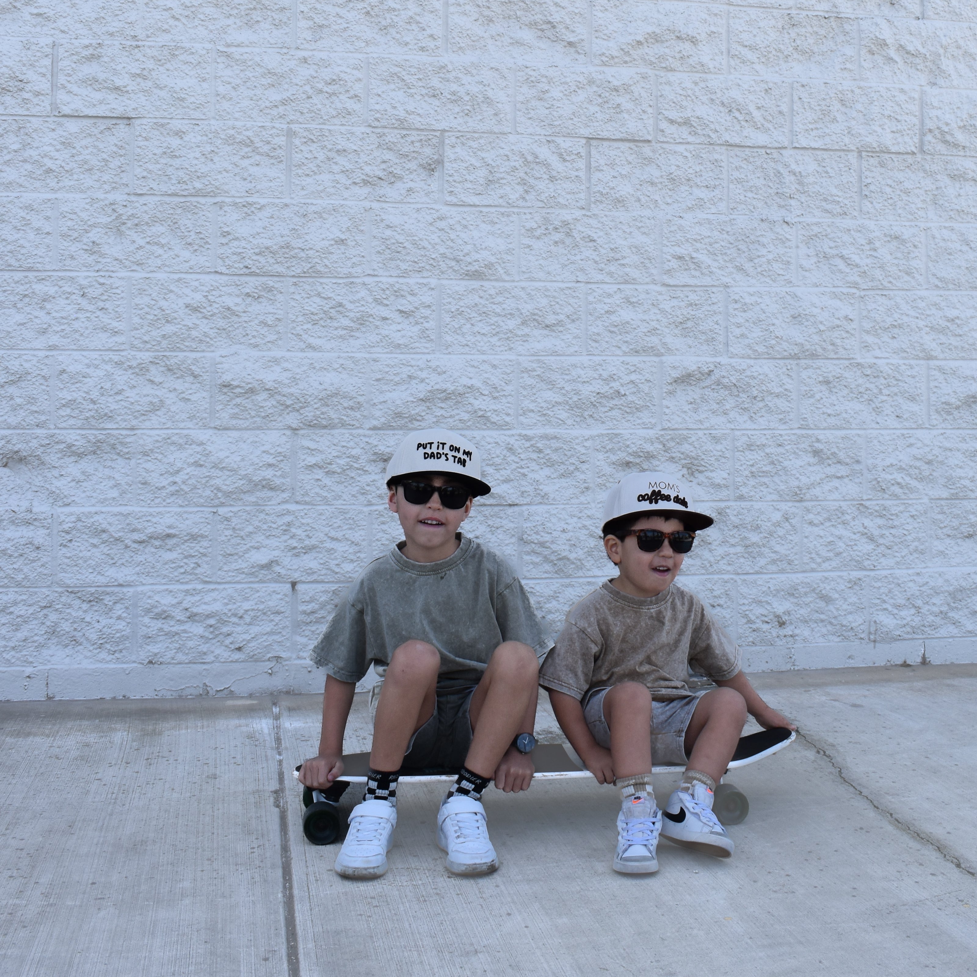 Two children sitting on a skateboard against a light-colored brick wall while wearing stone wash sets by rad toddler