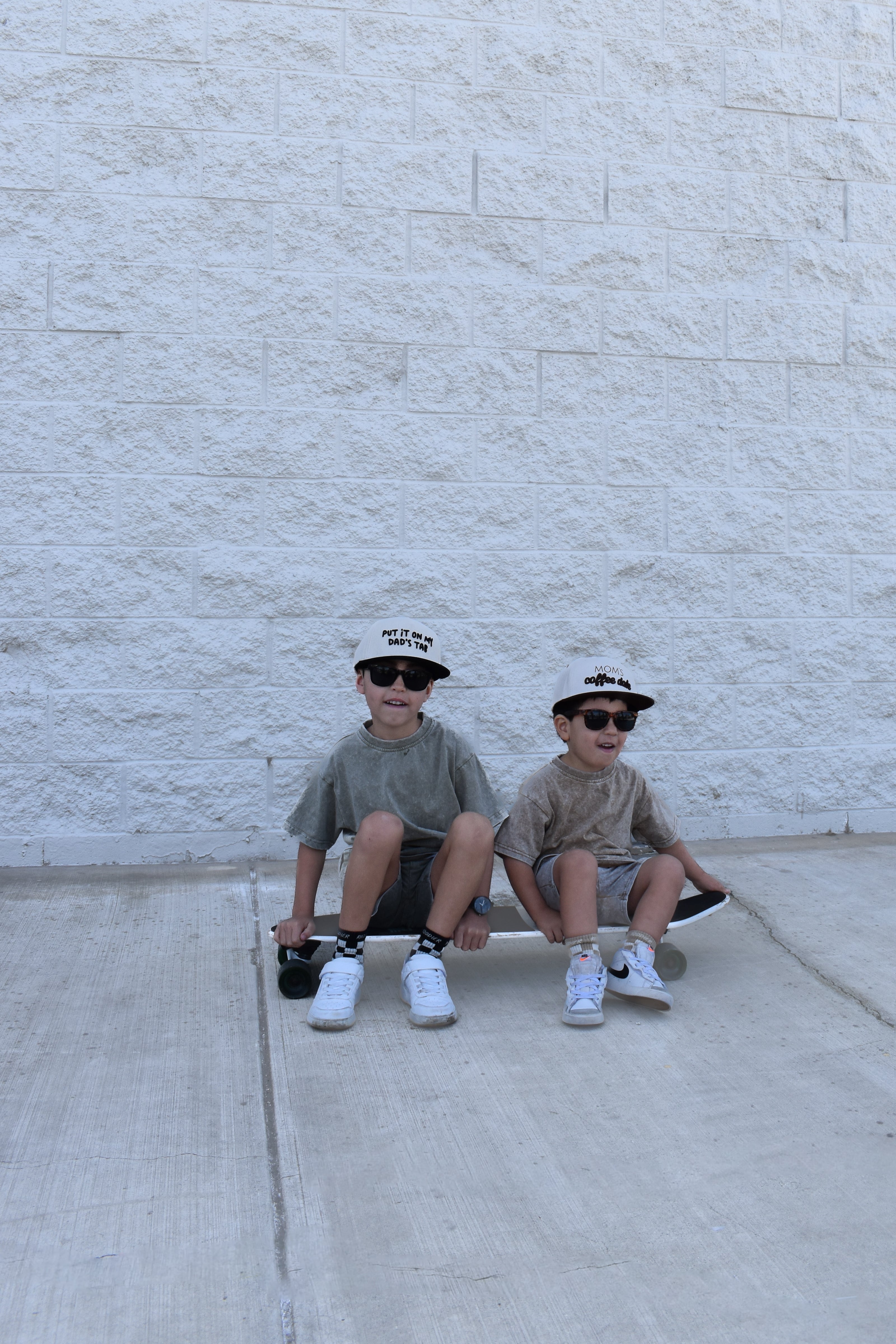 Two children sitting on a skateboard against a light-colored brick wall while wearing stone wash sets by rad toddler
