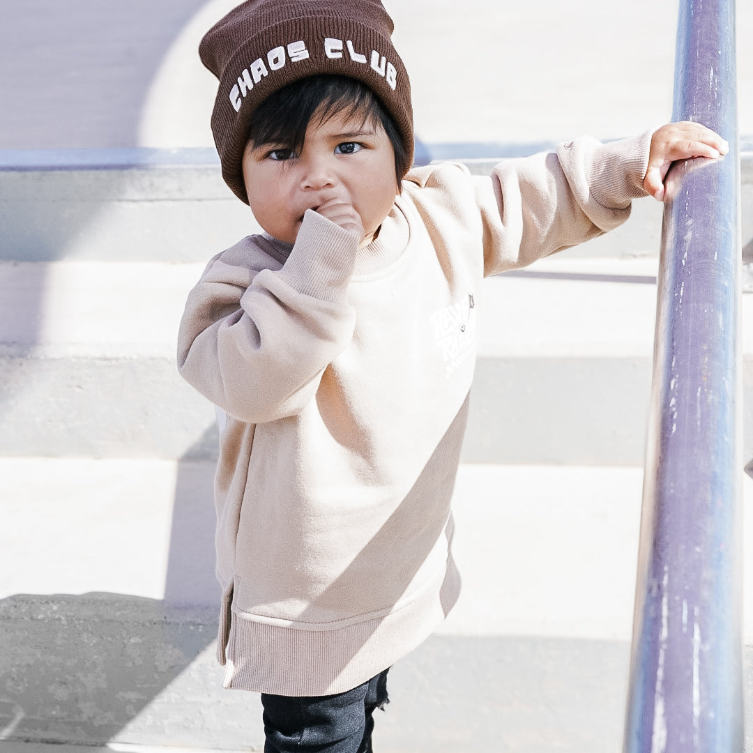 Child wearing a beige hoodie and black pants with a 'Chaos Club' beanie, standing on a playground.