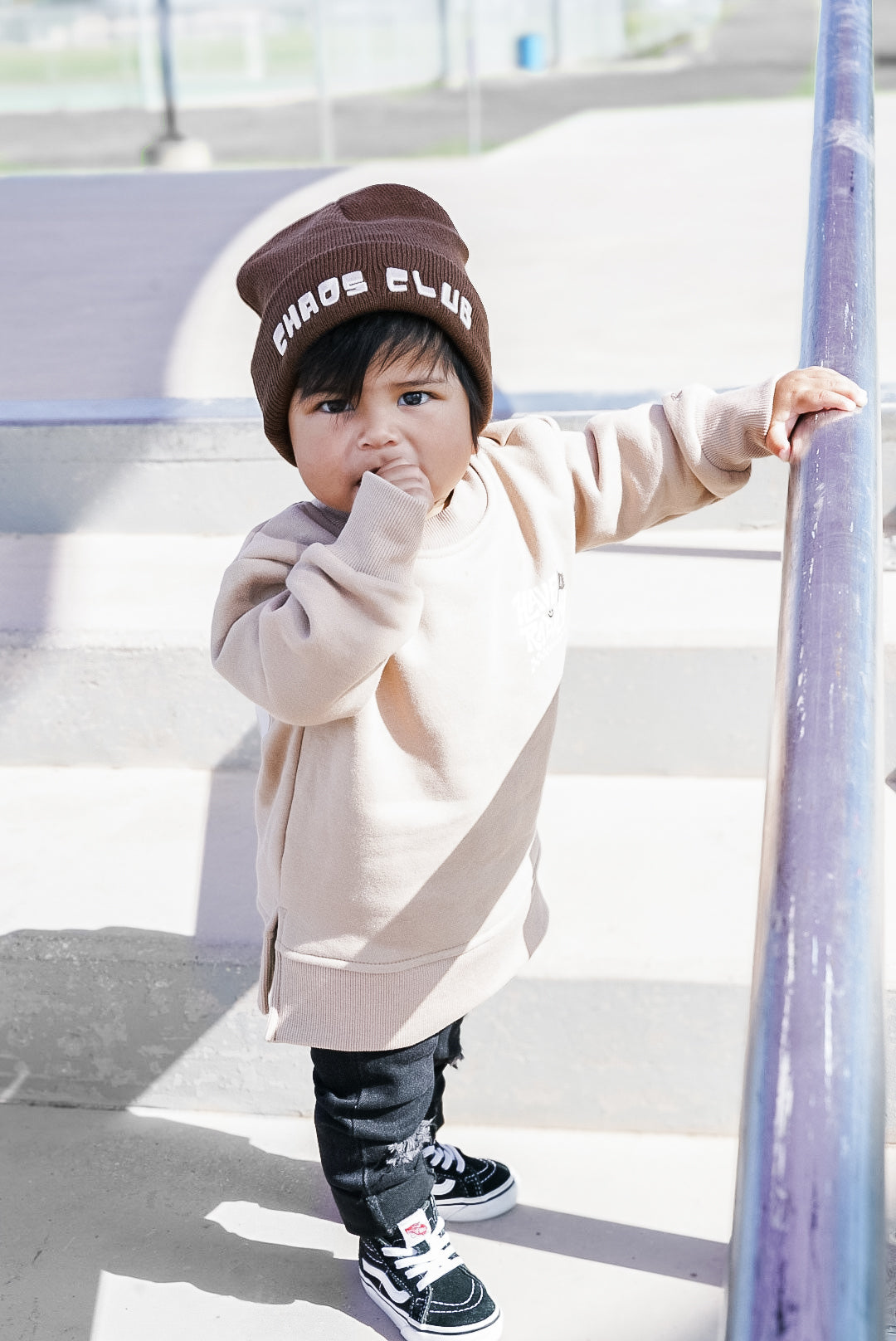 Child wearing a beige hoodie and black pants with a 'Chaos Club' beanie, standing on a playground.