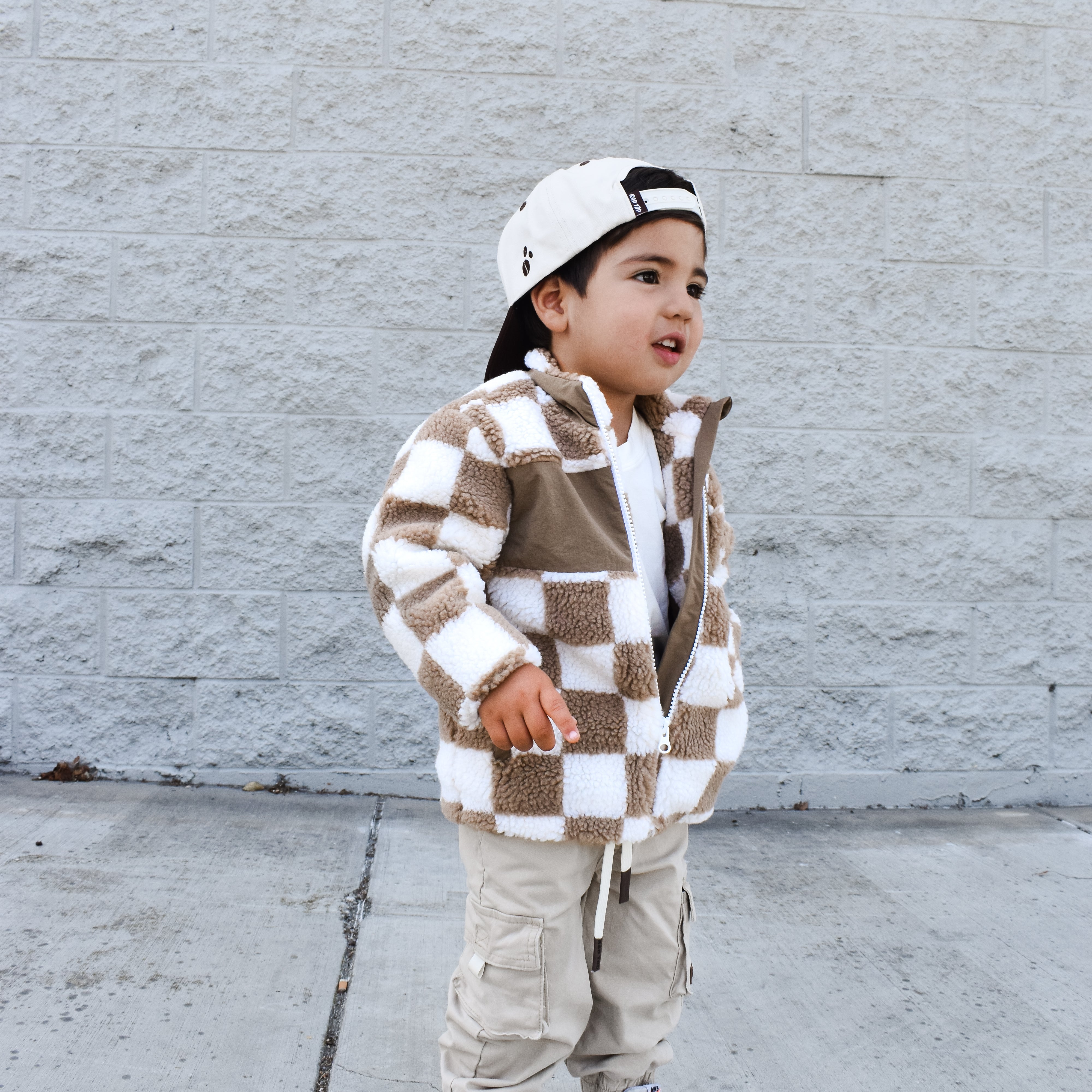 Child wearing a checkered jacket and cargo pants standing against a gray wall.