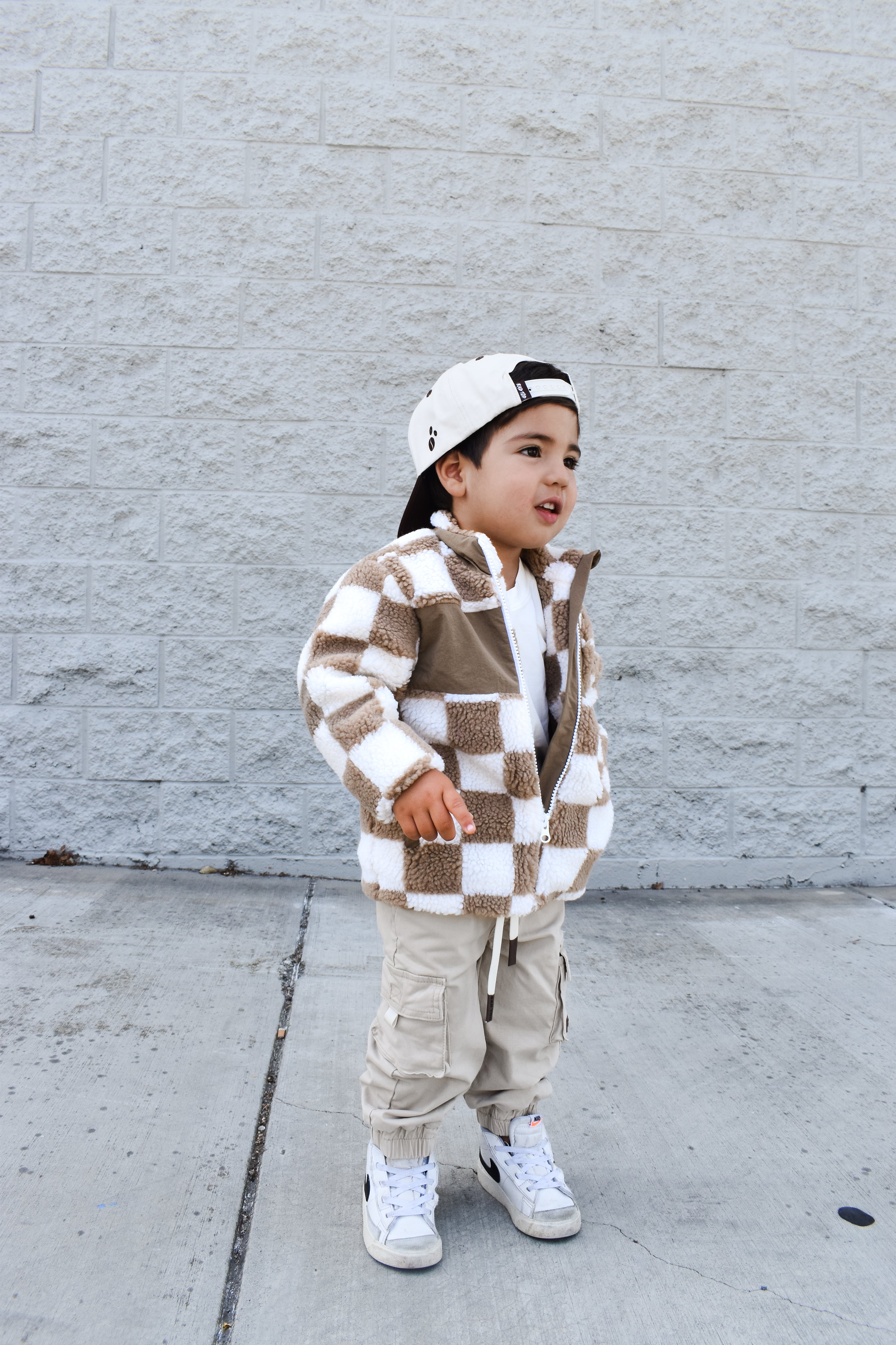 Child wearing a checkered jacket and cargo pants standing against a gray wall.