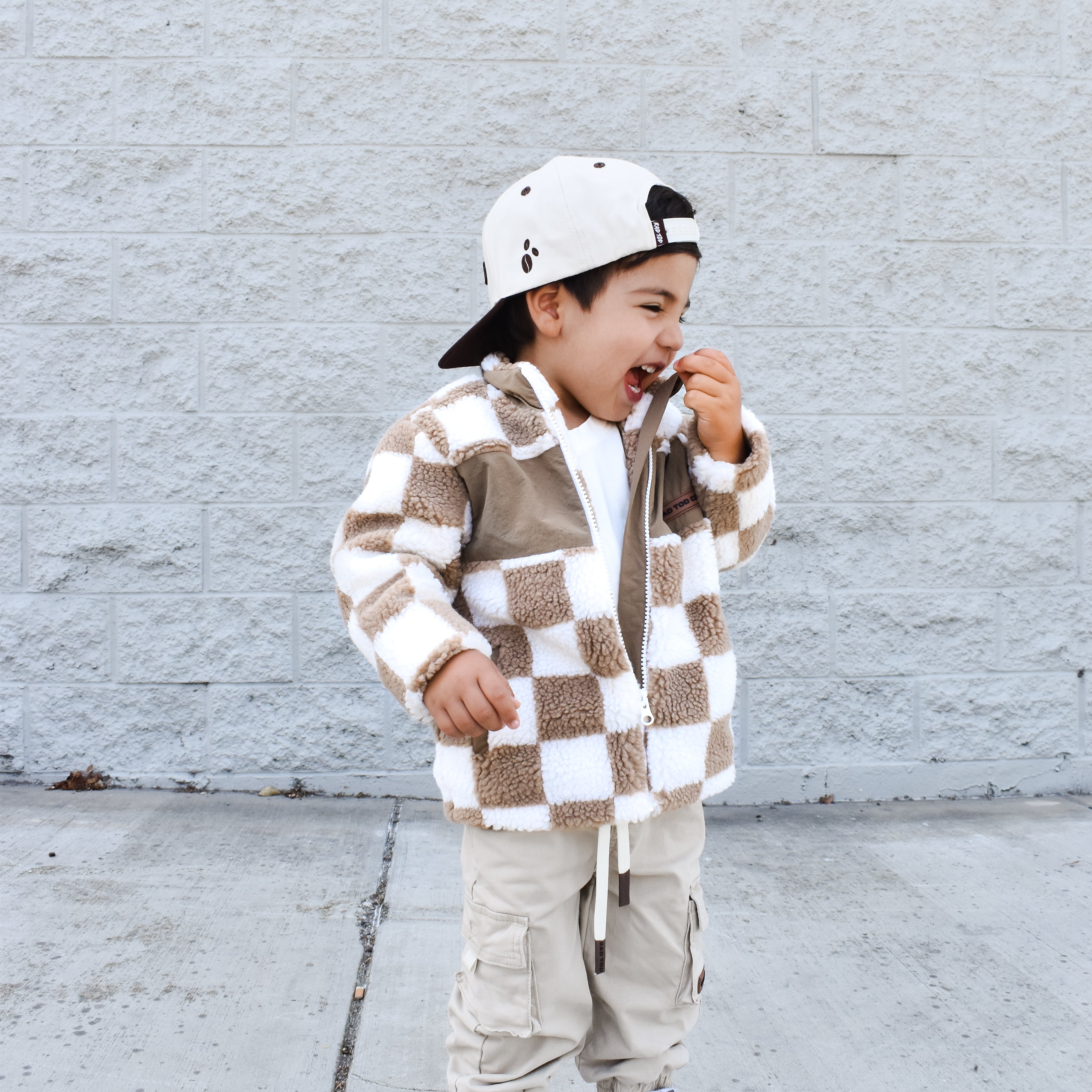 Child wearing a checkered jacket and cargo pants standing against a light gray brick wall.