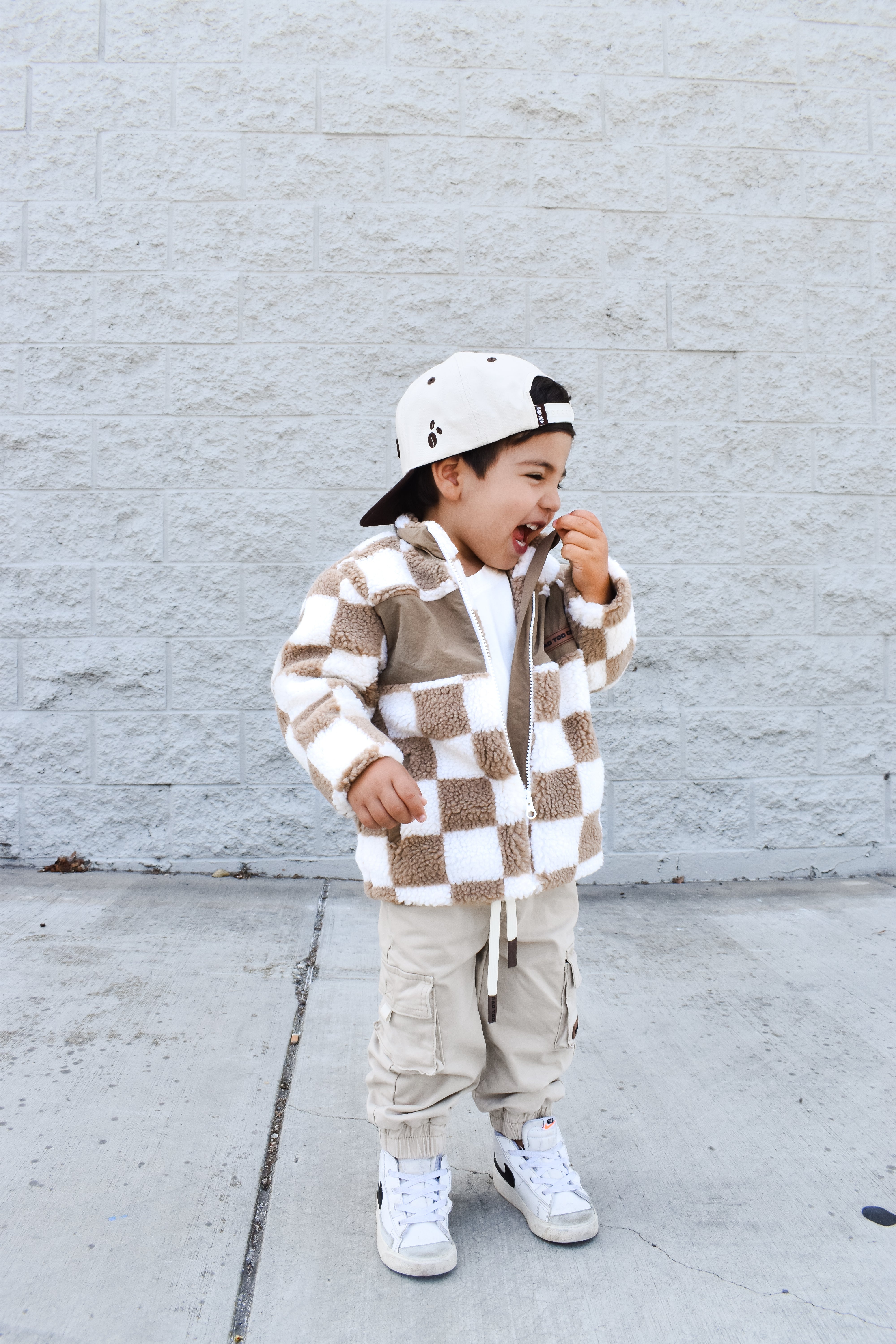 Child wearing a checkered jacket and cargo pants standing against a light gray brick wall.