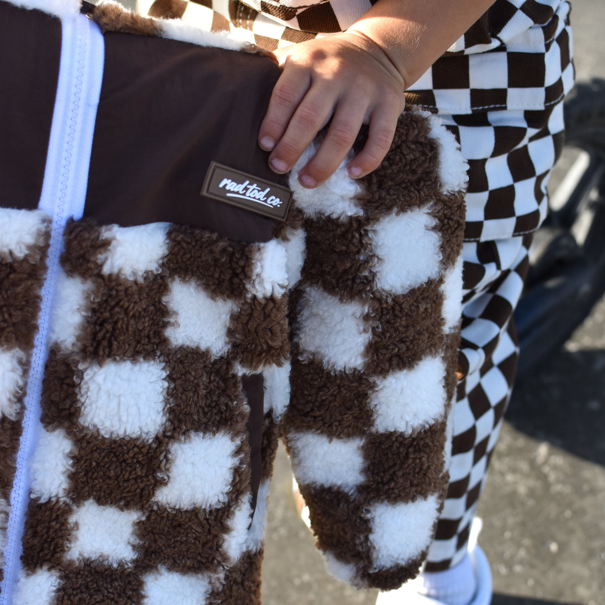 A close-up of a person holding a brown and white checkered teddy sherpa jacket outdoors. The person is wearing matching brown and white checkered pants, white slip-on shoes, and a gray shirt