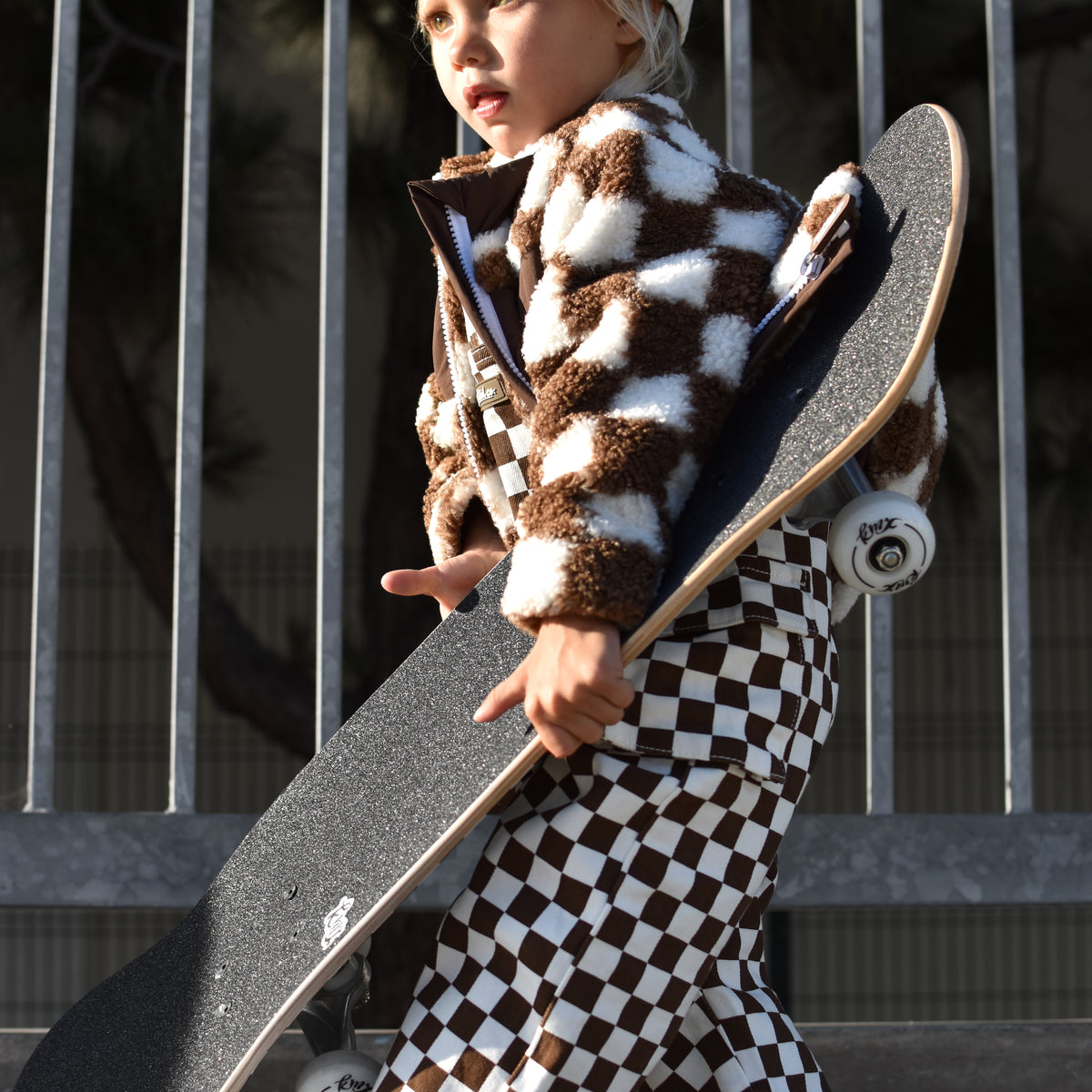 A toddler walking while carrying a skateboard, wearing a brown and white checkered fuzzy jacket and matching checkered pants, a white cap, and white sneakers.