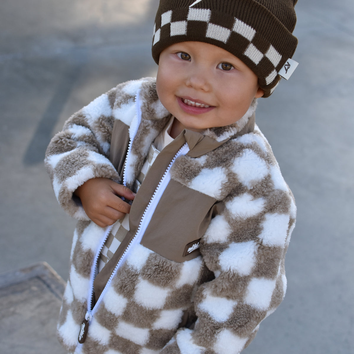 A smiling toddler wearing a brown and white checkered fuzzy zip-up jacket, matching checkered pants, and a brown beanie with a white lightning bolt, standing outdoors on a concrete surface.
