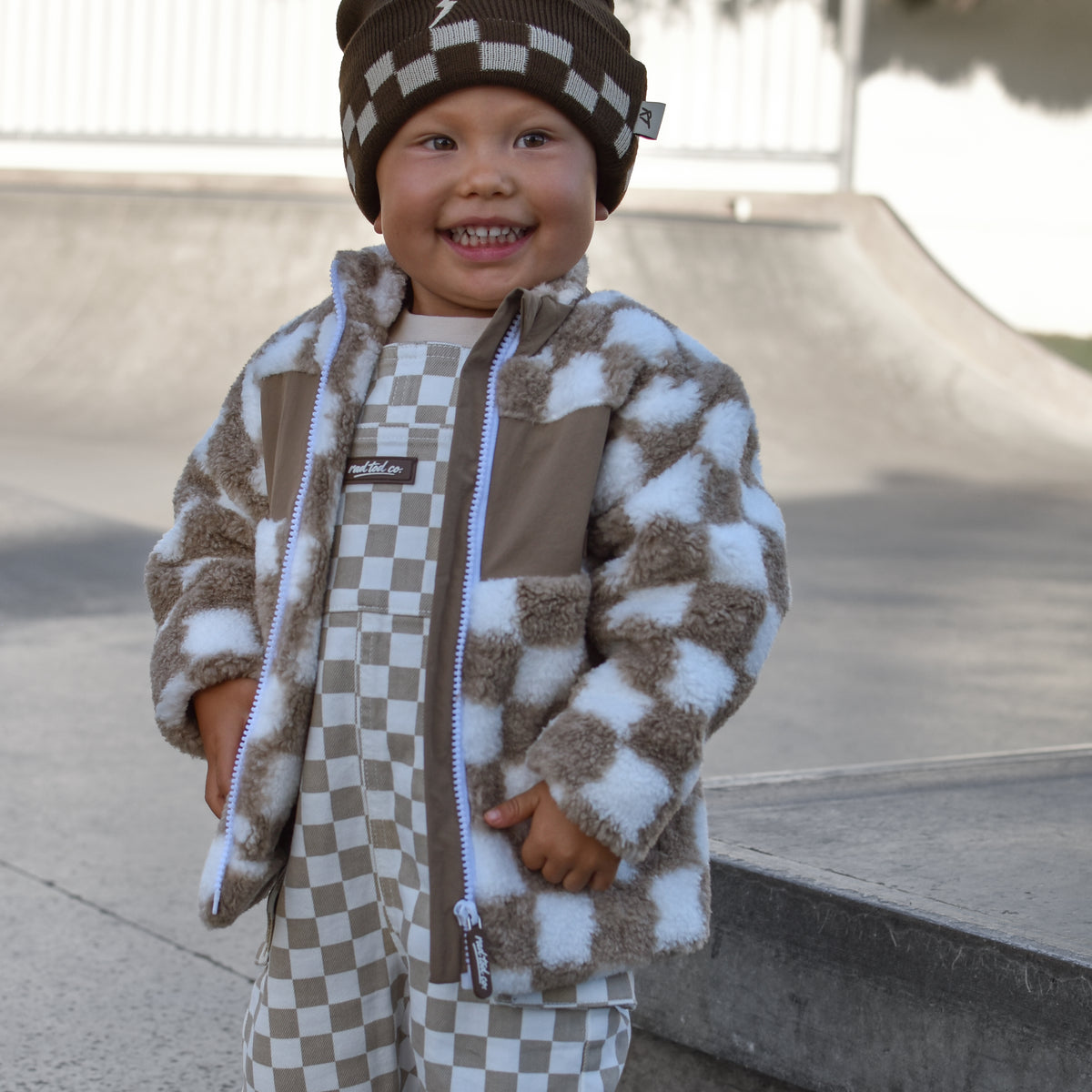 A smiling toddler standing outdoors in a skatepark, wearing a brown and beige checkered fuzzy jacket, matching checkered pants, and a brown beanie with a checkerboard pattern.