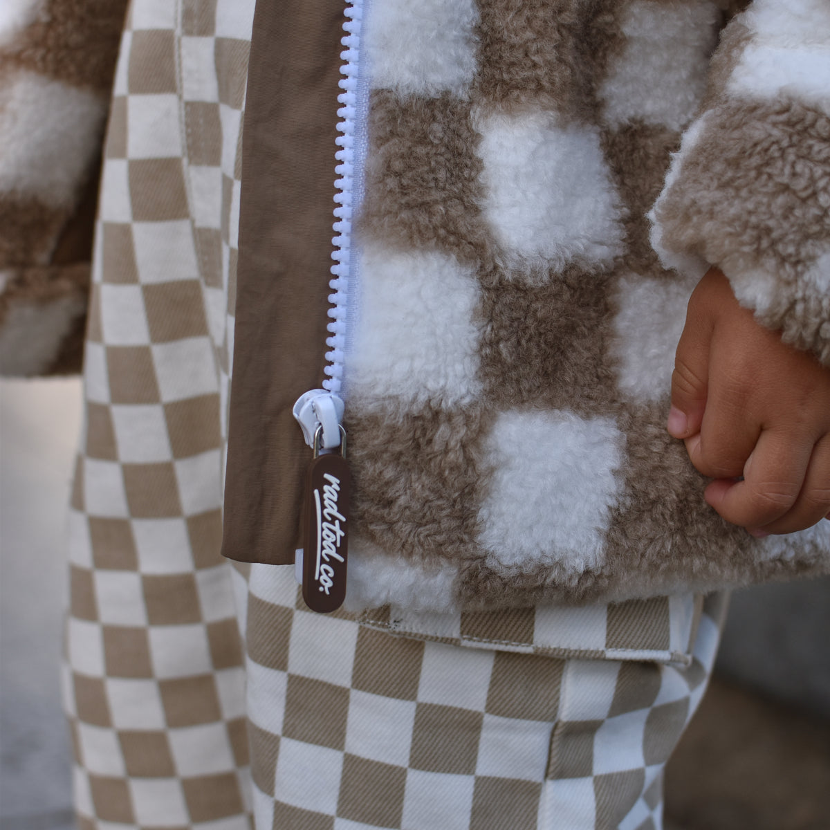 Close-up of a child wearing a brown and cream checkered fleece zip-up jacket and matching checkered pants, with a hand visible at the side.