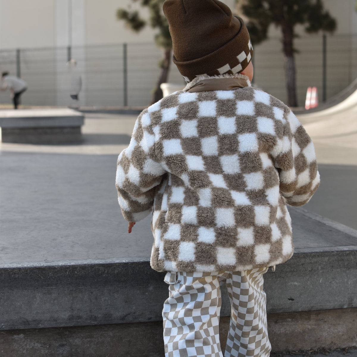 A toddler seen from behind wearing a brown and white checkered fuzzy jacket and matching pants, walking in an outdoor concrete plaza.