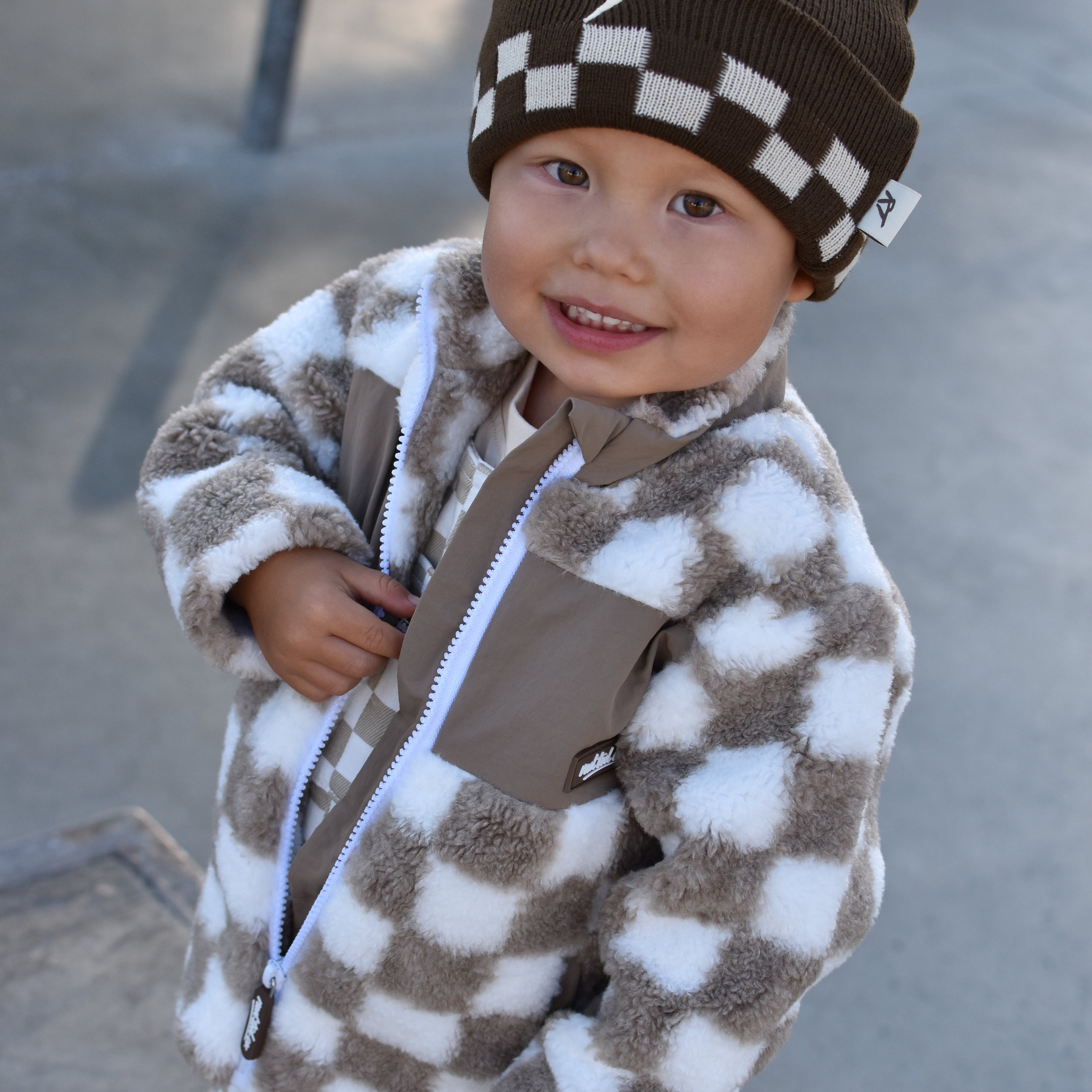 A smiling toddler wearing a brown and white checkered fuzzy zip-up jacket, matching checkered pants, and a brown beanie with a white lightning-bolt patch, photographed outdoors at a skatepark.