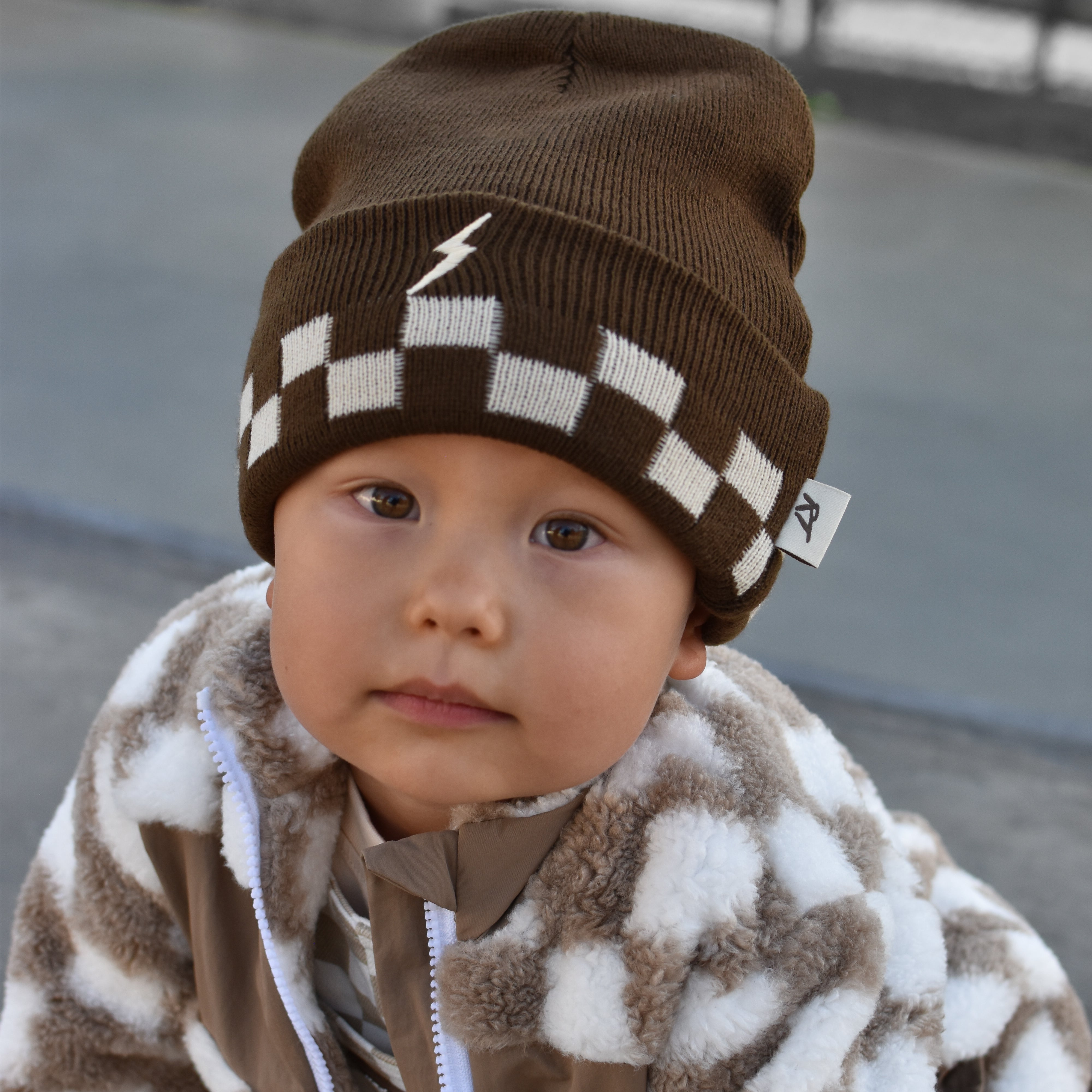 A baby wearing a brown and white checkered knit beanie with a small white lightning bolt logo, paired with a brown and white patterned fleece jacket, photographed outdoors.