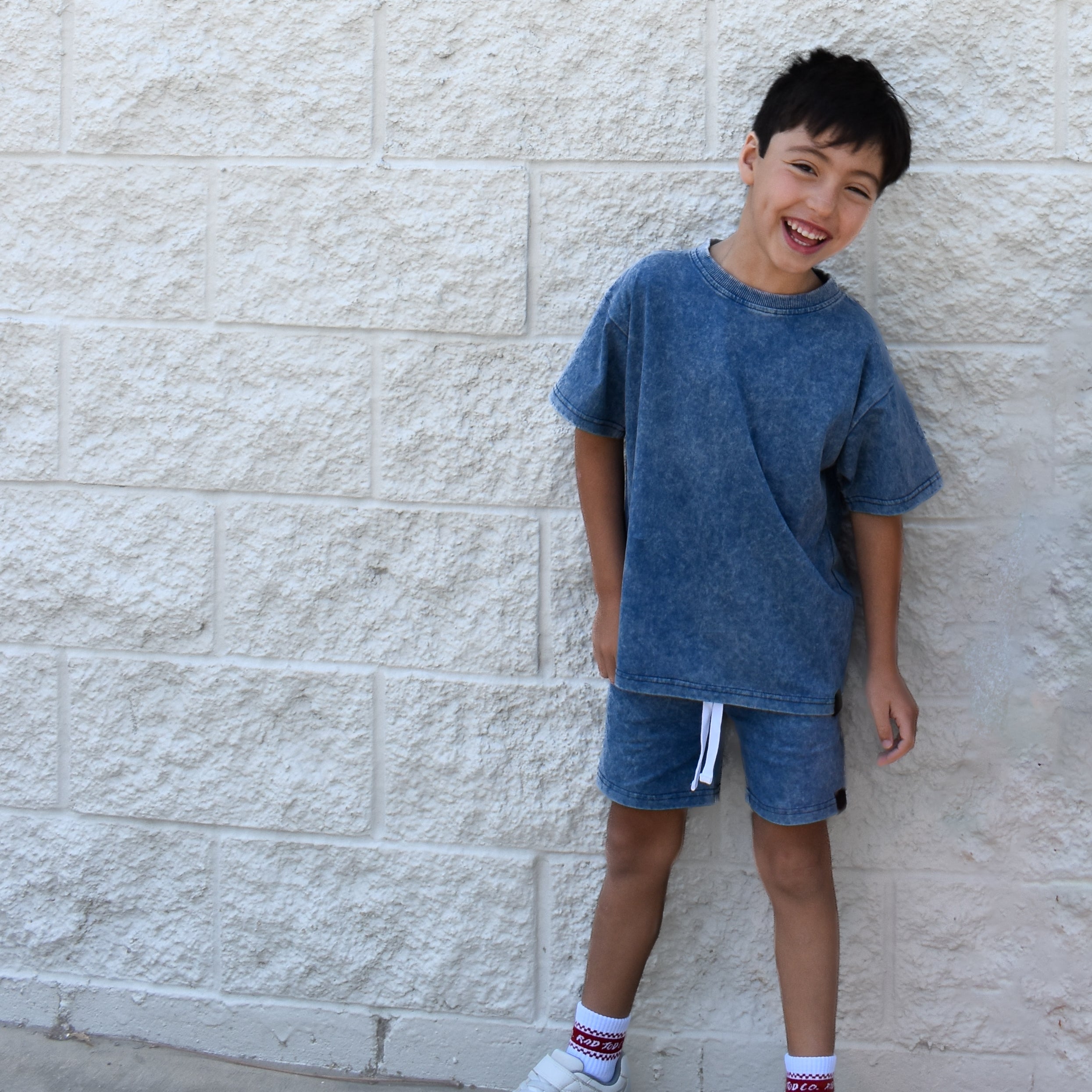 Child wearing a blue t-shirt and shorts standing against a light stone wall.