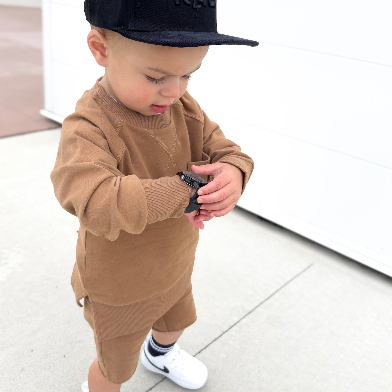 Young boy wearing almond Core Collection toddler set, standing outdoors and showcasing the relaxed, above-the-knee fit checking the time!