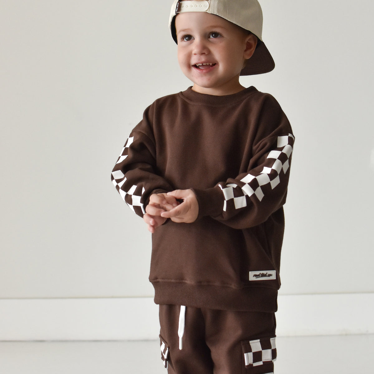 A toddler wearing a matching brown sweatshirt and jogger pants set with black and white checkered sleeve and leg details, along with a beige baseball cap, standing against a plain light gray wall.