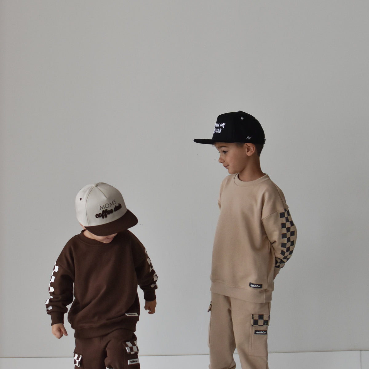 Two children modeling matching streetwear tracksuits in a minimalist studio setting. The child on the left wears a dark brown long-sleeve sweatshirt and matching pants with white graphic details and a white cap.