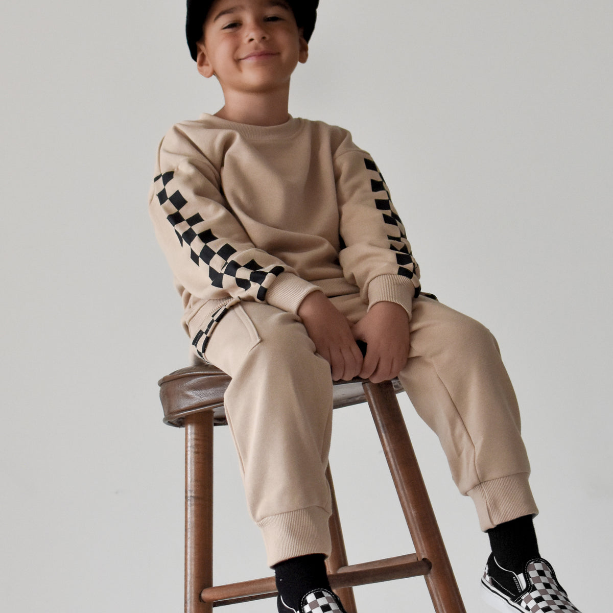A young boy wearing a beige sweatshirt and jogger pants with black-and-white checkerboard sleeve and sock details, sitting on a wooden stool against a light gray background.