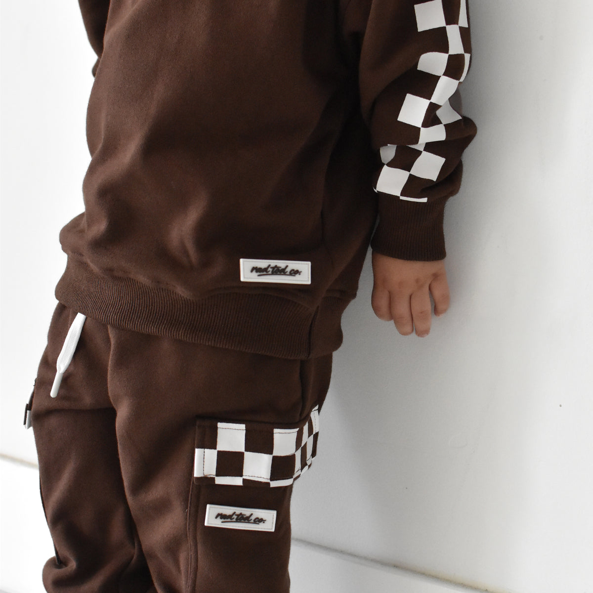 A child wearing a matching brown sweatshirt and jogger pants set with white and black checkerboard accents on the sleeves and cargo pockets, standing against a white wall.
