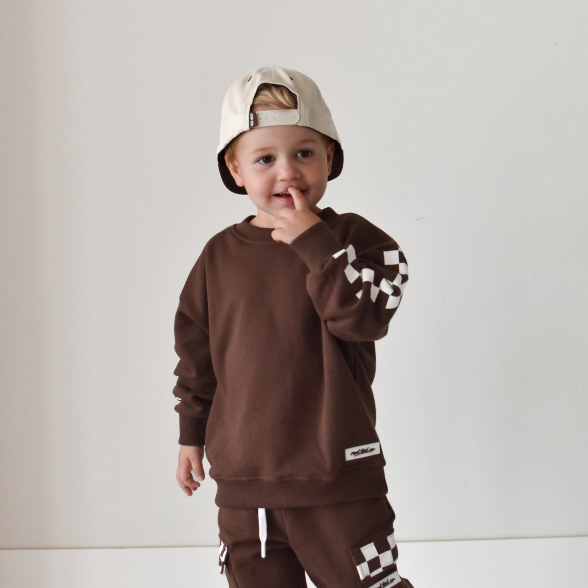 A toddler wearing a matching brown sweatshirt and jogger pants set with white checkerboard accents, a beige baseball cap, and black sneakers, standing against a plain white wall.