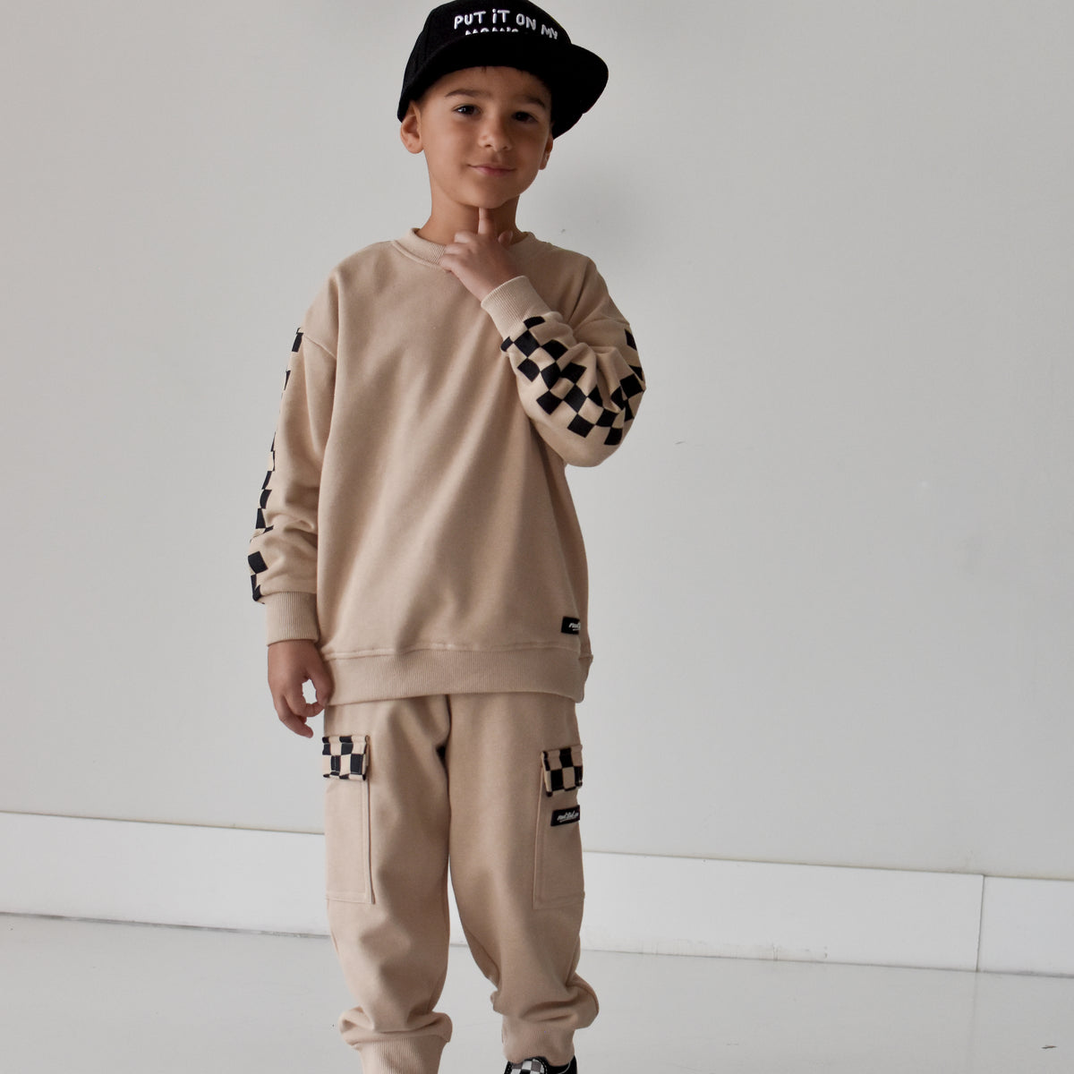 A young boy modeling a beige two-piece tracksuit with black and white checkerboard pattern details on the sleeves, pants, and cap, standing against a plain white studio background.