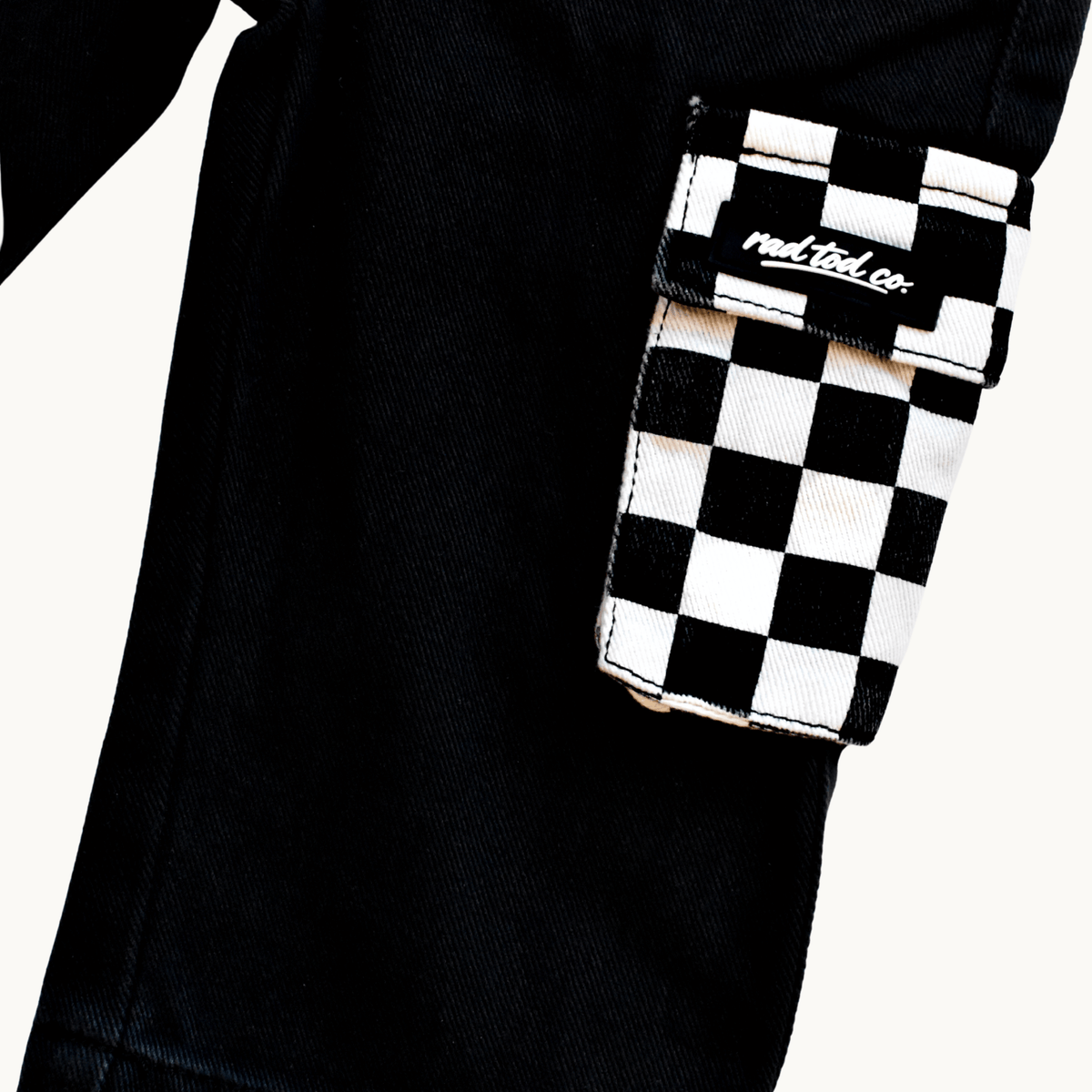 Close-up of black denim pants with a flap pocket featuring a black and white checkerboard pattern and a visible 'Rad Tod Co.' logo patch.