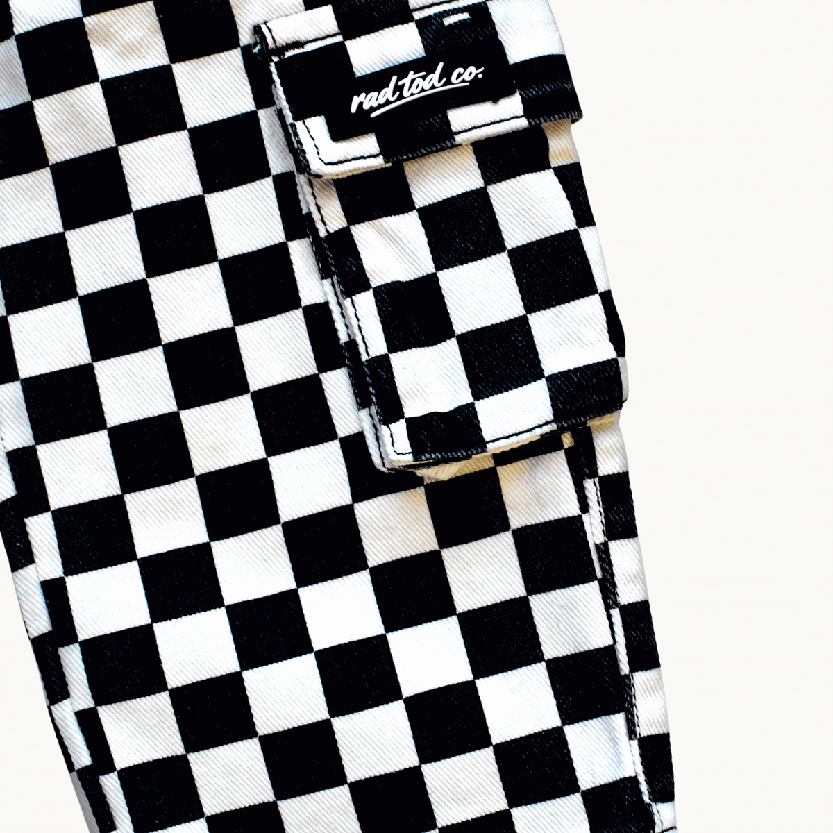 Close-up of a black and white checkered cargo pocket with a flap and a visible 'Rad Tod CO.' label, on a light off-white background.