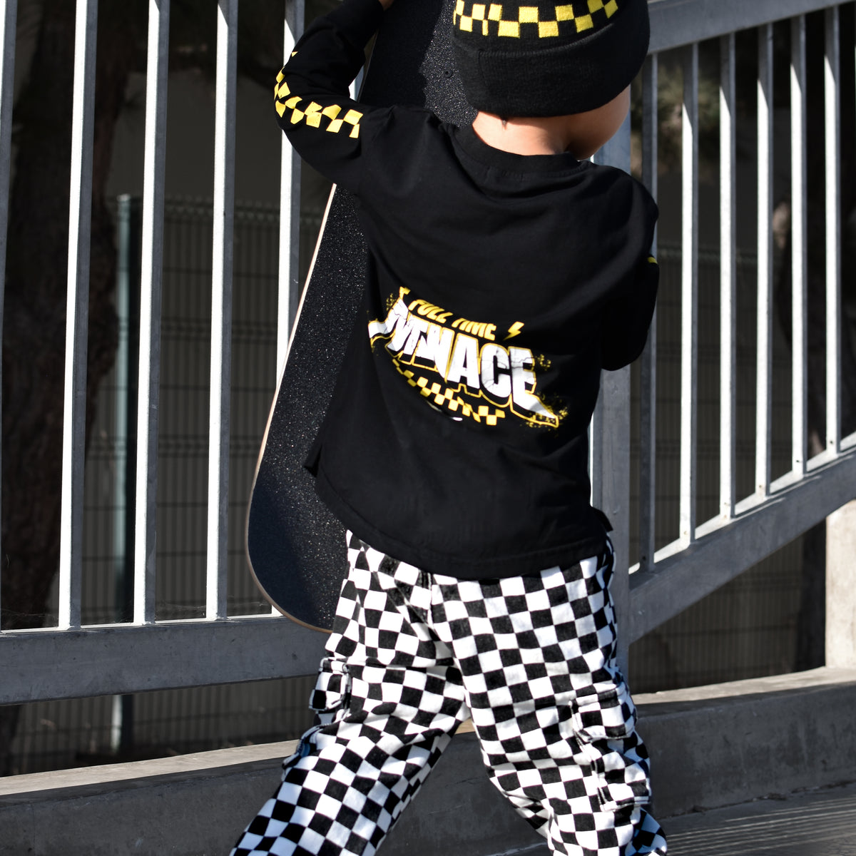 A child wearing a black and yellow checkered beanie, a black long-sleeve top with a yellow and white 'MENACE' graphic on the back, and black-and-white checkered pants, holding a skateboard outdoors near a metal railing.