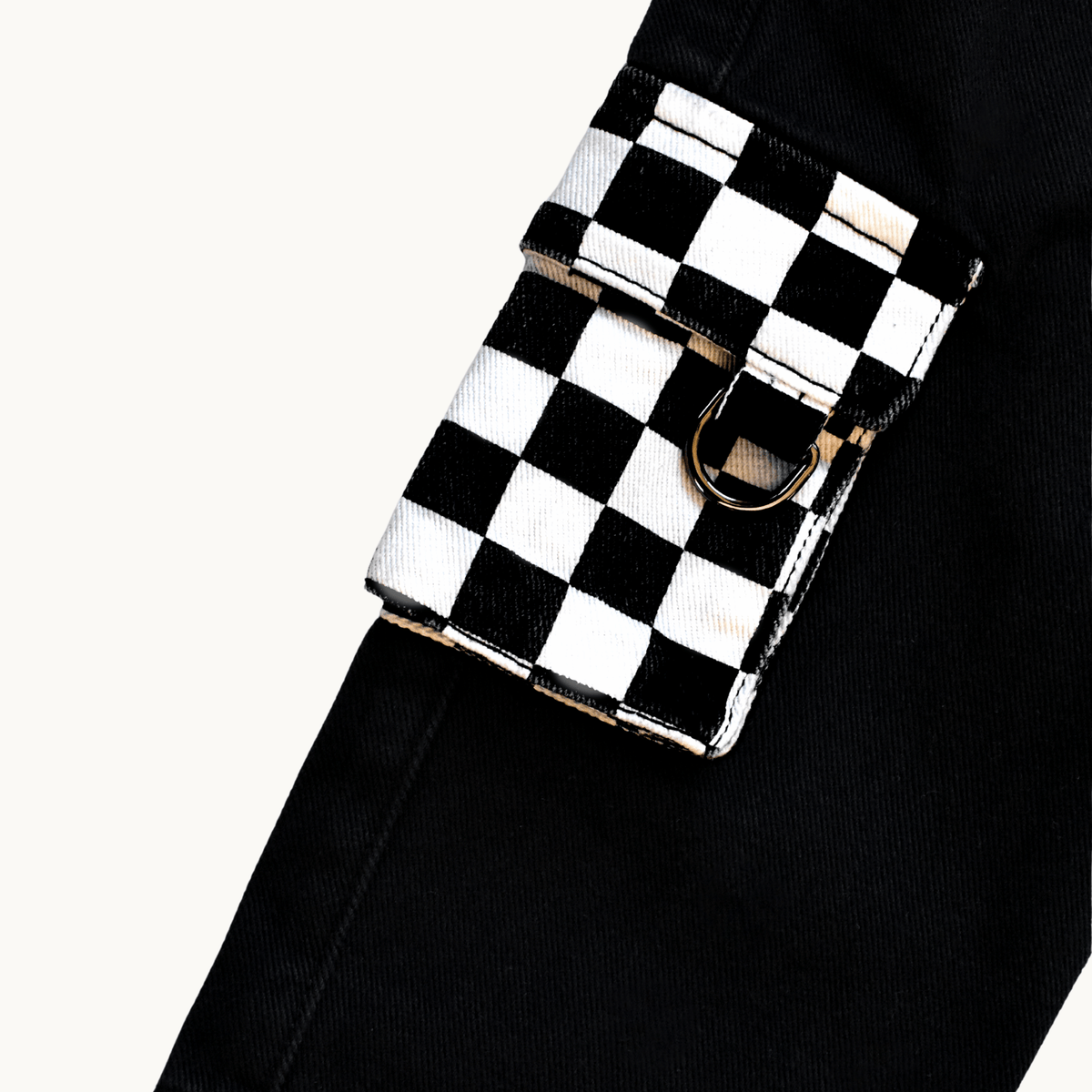 A close-up of a black and white checkerboard flap pouch with a silver-tone metal D-ring, attached to a black denim belt or pants leg, set against a plain off-white background.