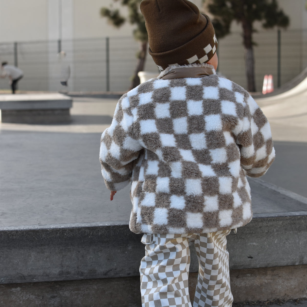A toddler seen from behind wearing a brown and white checkerboard fleece jacket and matching pants, walking outdoors in a skatepark setting.