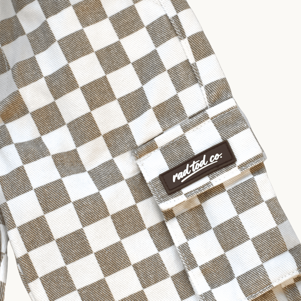 Close-up of a checkered cargo pocket in a brown and white checkerboard pattern, featuring a black brand patch that reads 'Rad Tod co.' on a light off-white background.