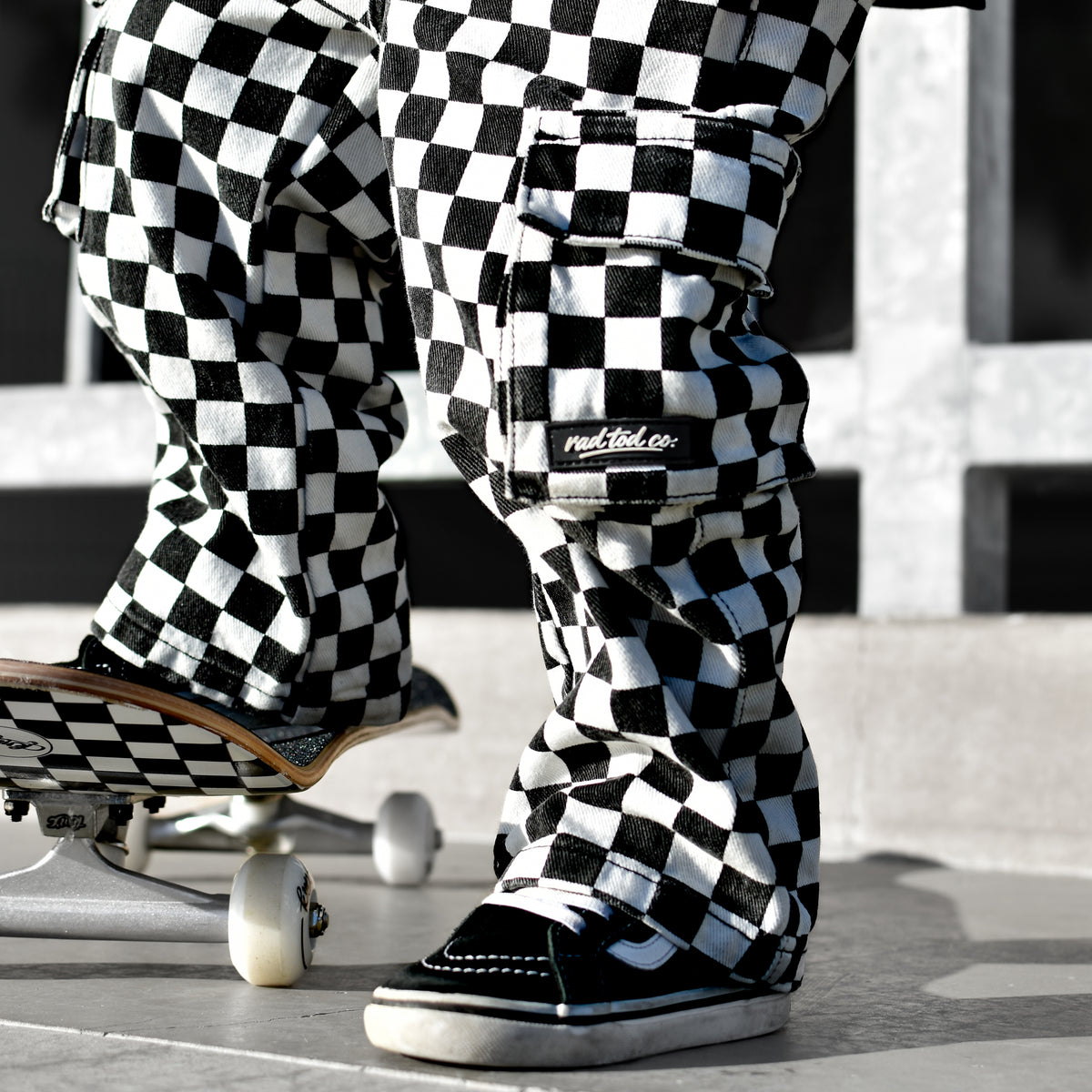 A person wearing black and white checkered cargo pants and black slip-on shoes stands on concrete next to a skateboard. The pants have a bold black-and-white checkerboard pattern and a visible 'Rad Tod CO. ' brand patch on the pocket.
