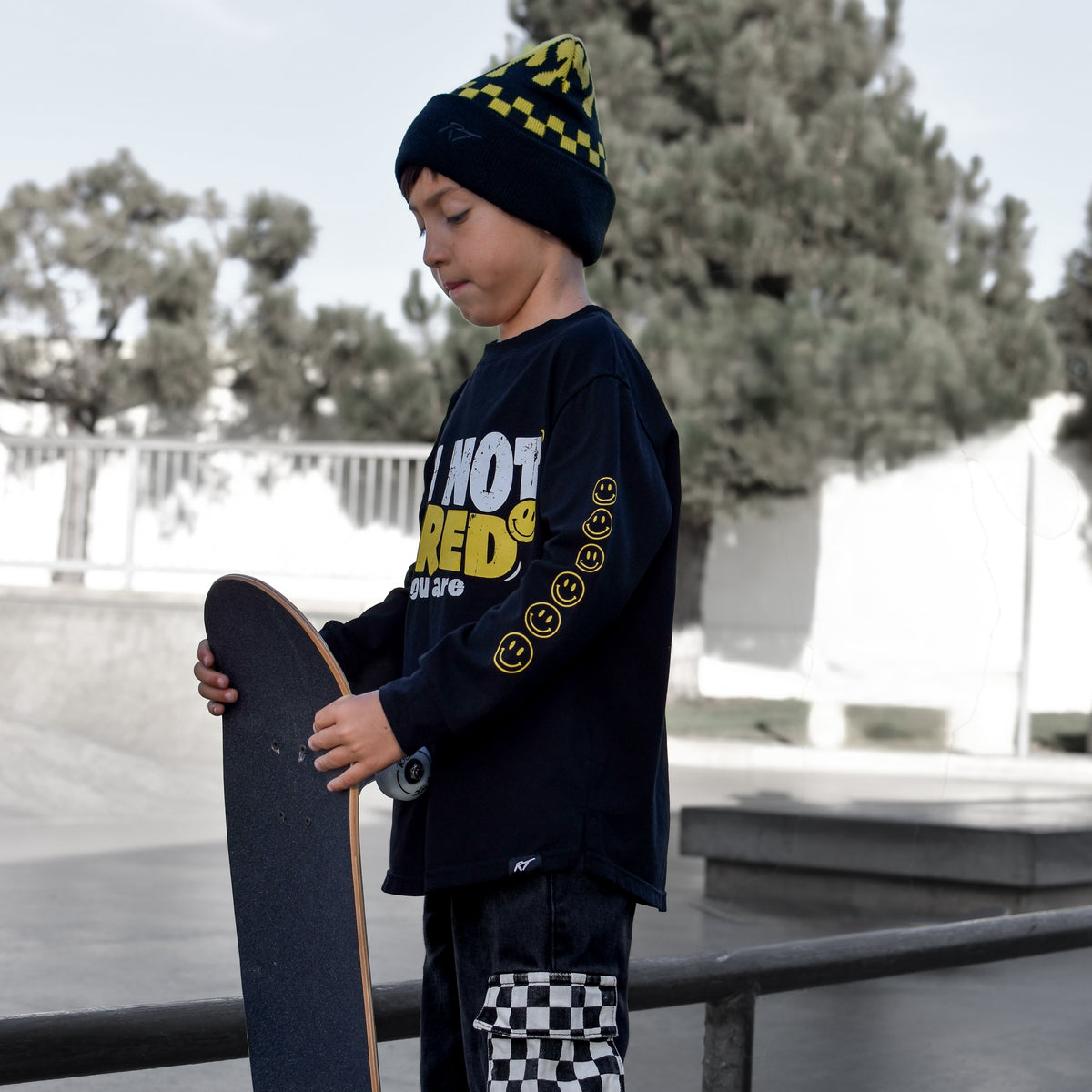 A child standing at a skatepark holding a skateboard, wearing a black beanie with yellow checkered trim, a black long-sleeve graphic shirt with yellow text, and black pants with a checkered patch on the lower leg.