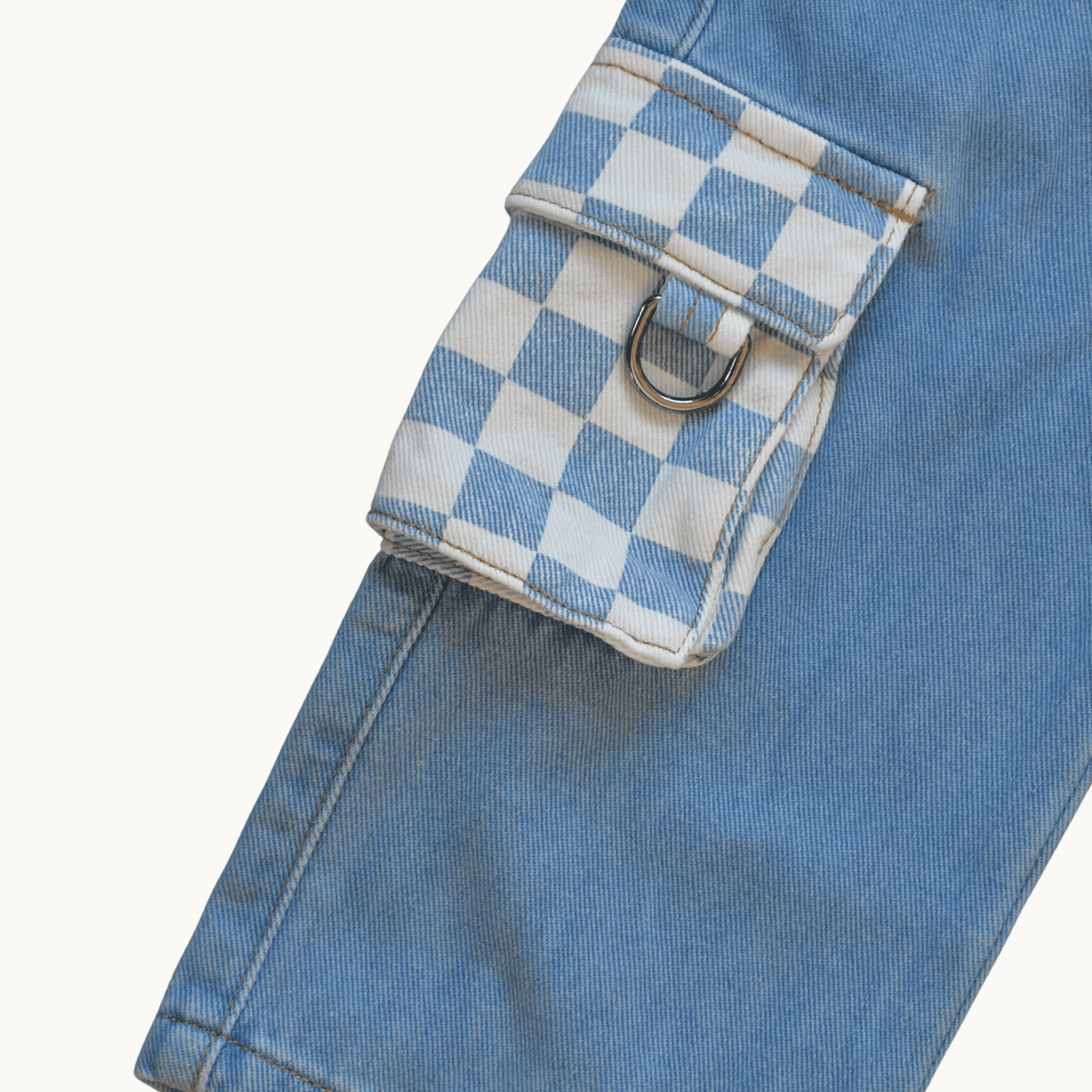Close-up of a light blue denim jacket or pants leg showing a flap cargo pocket with a white and blue checkered pattern and a silver metal D-ring, on a plain off-white background.