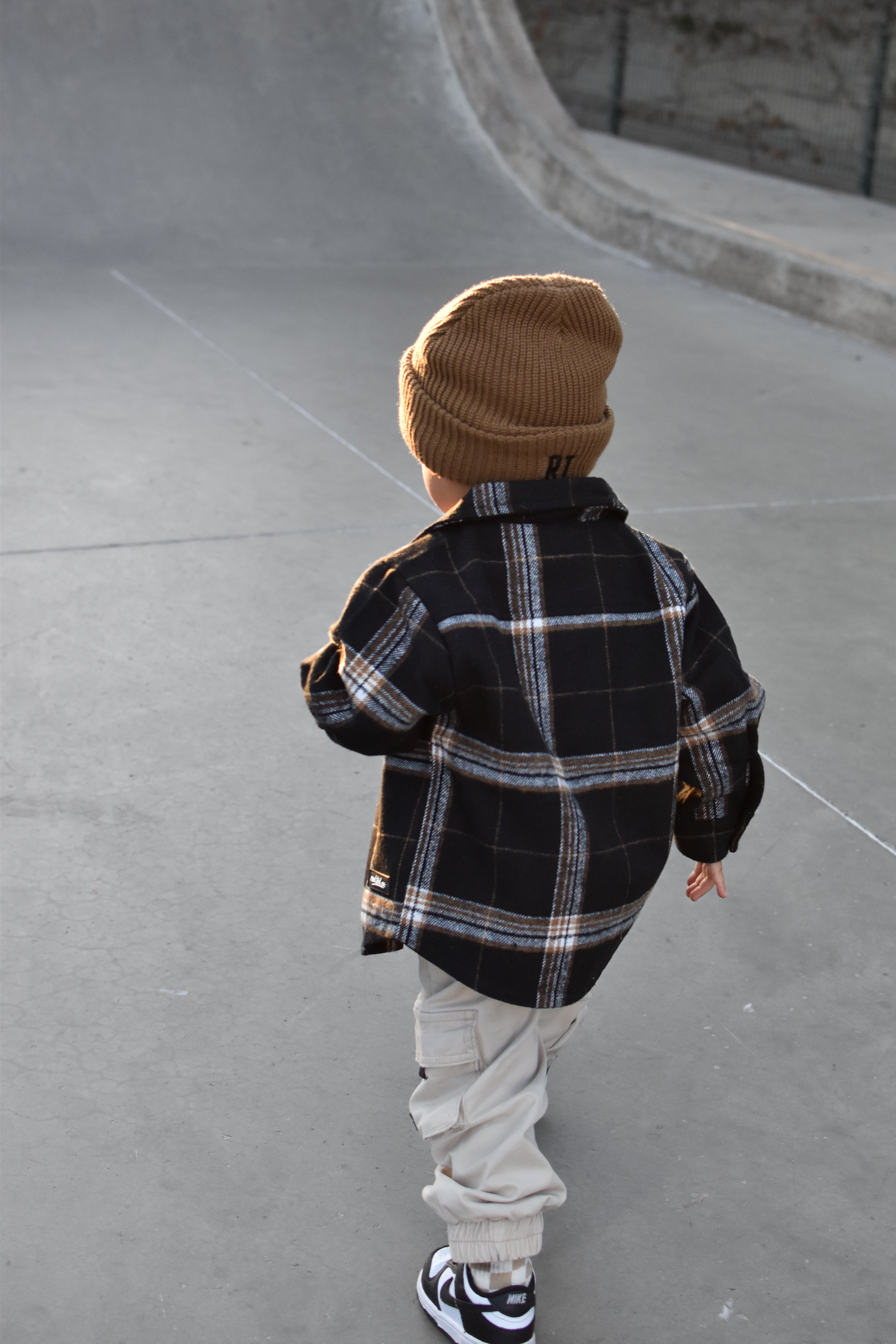 A toddler viewed from behind wearing a tan knit beanie, a black and white plaid flannel shirt, light beige pants, and black and white sneakers, standing on a concrete skatepark surface.