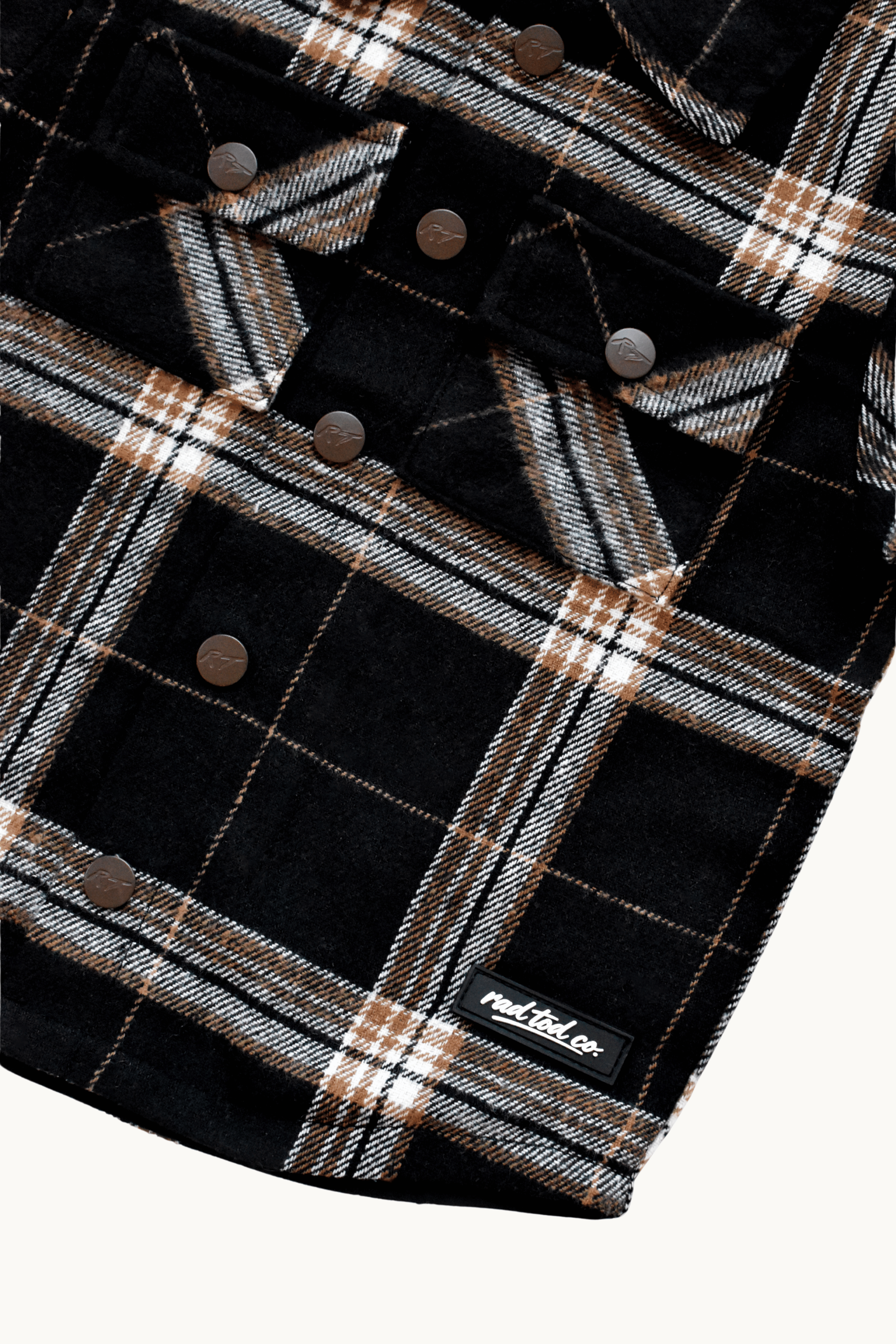 Close-up of a black plaid flannel shirt jacket with brown buttons and a small 'RAD TOD co.' label near the bottom right, shown on a light background.