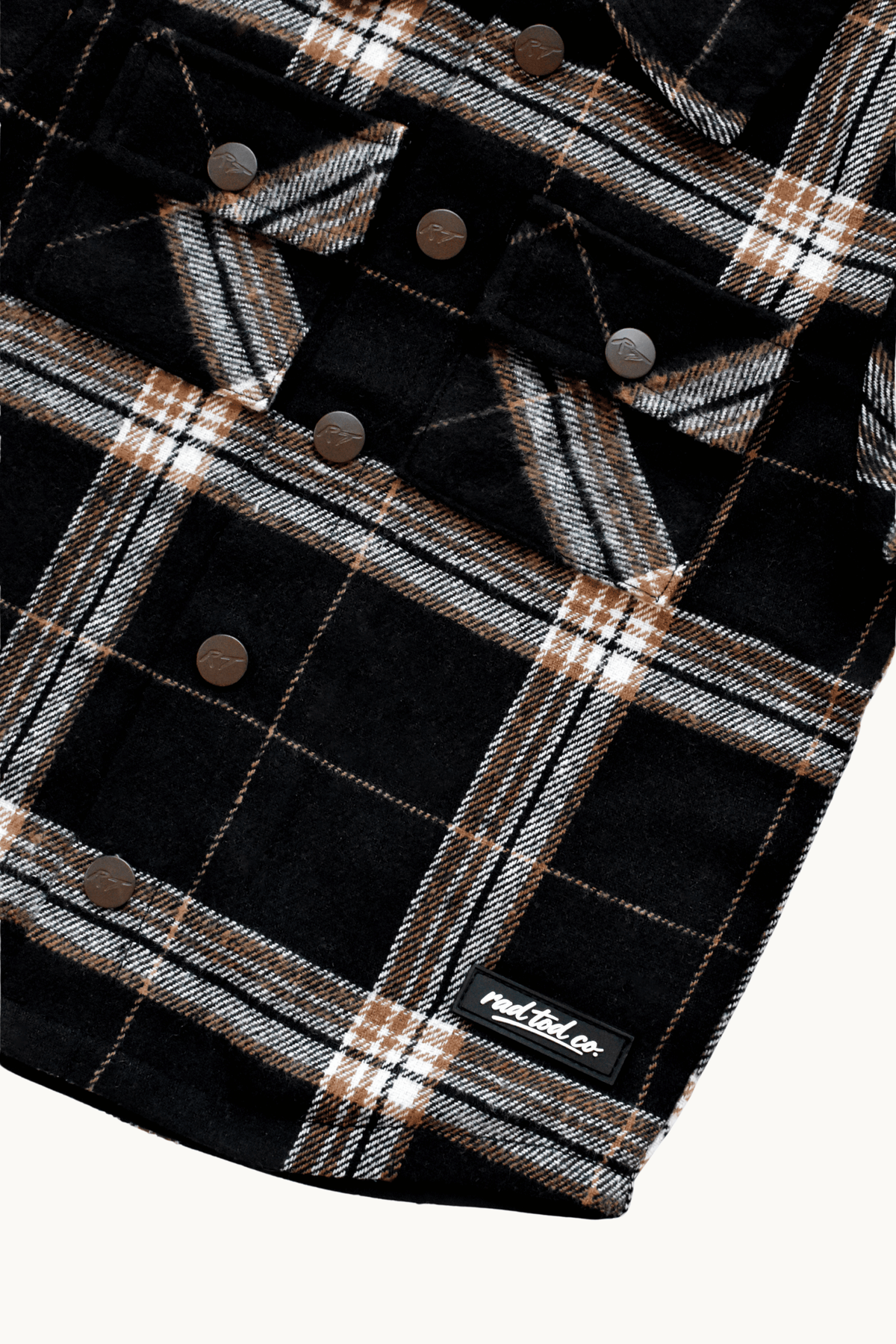 Close-up of a black plaid flannel shirt jacket with brown buttons and a small 'RAD TOD co.' label near the bottom right, shown on a light background.
