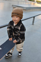 A toddler standing at a skatepark holding a skateboard, wearing a brown knit beanie with black text, a black and brown plaid flannel shirt, beige cargo pants, and black and white sneakers.