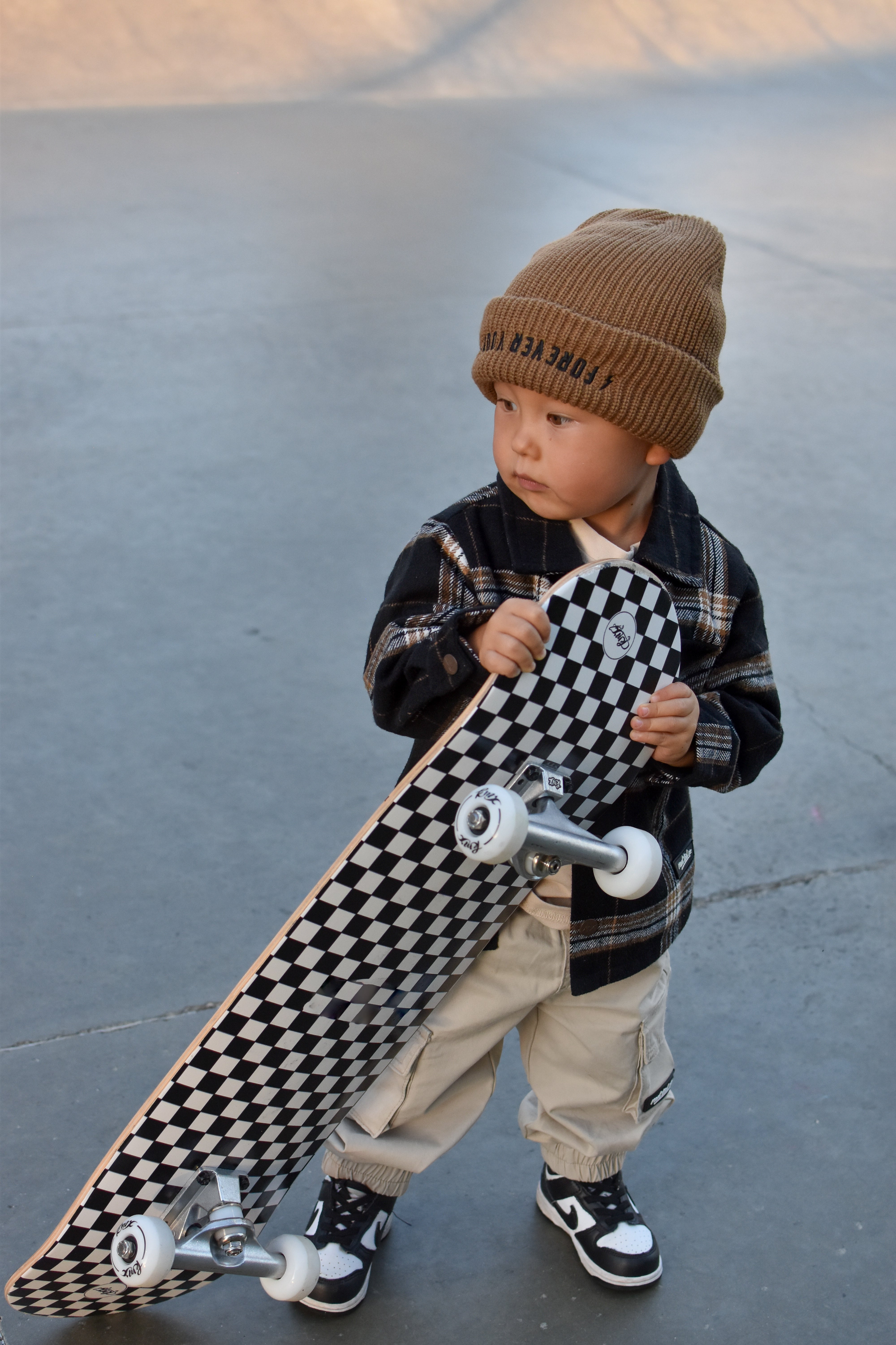 A toddler stands outdoors holding a skateboard with a black-and-white checkerboard deck. The child wears a tan knit beanie with dark lettering, a dark plaid jacket, light beige pants, and black-and-white sneakers.
