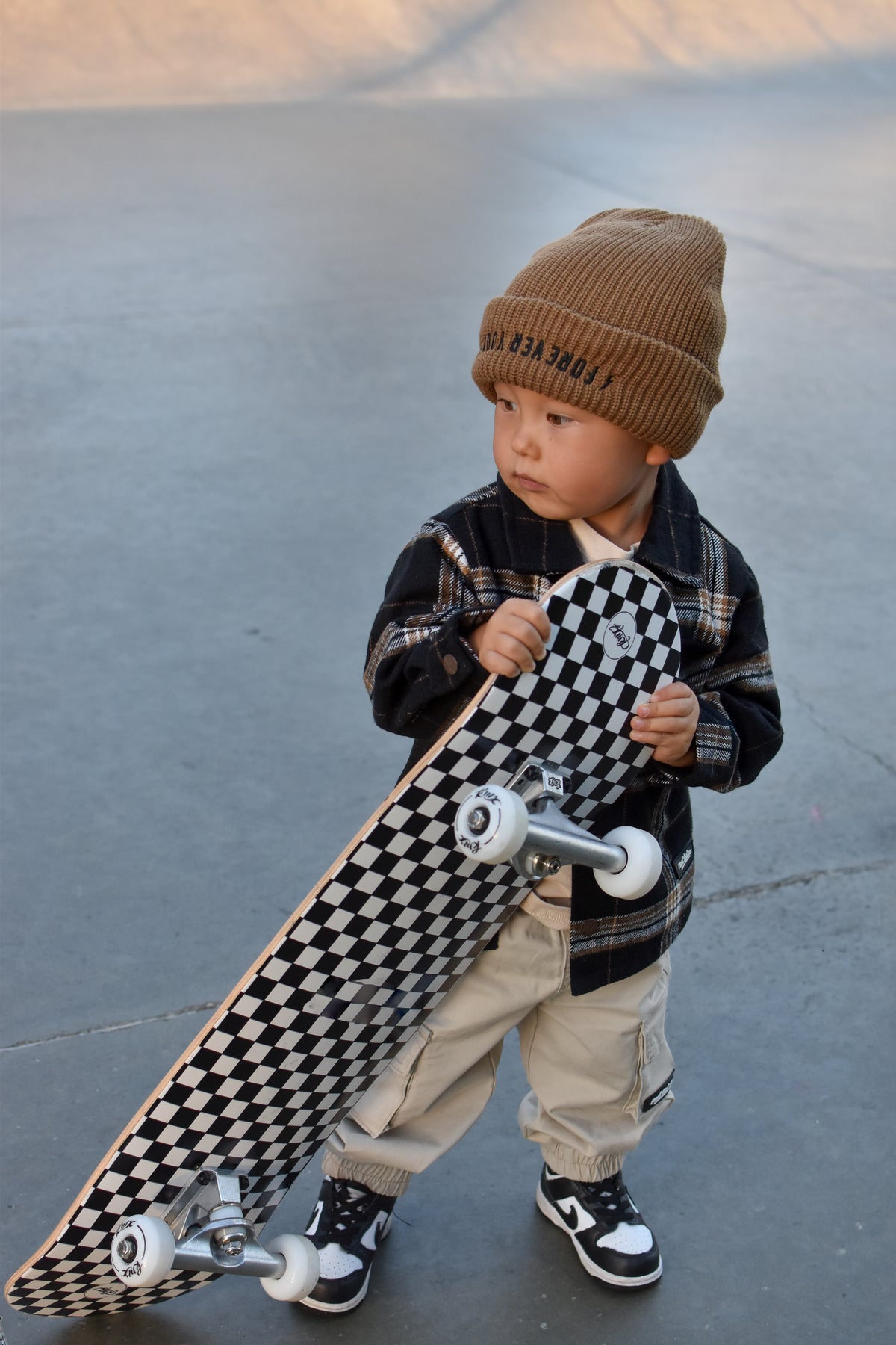 A toddler stands outdoors holding a skateboard with a black-and-white checkerboard deck. The child wears a tan knit beanie with dark lettering, a dark plaid jacket, light beige pants, and black-and-white sneakers.