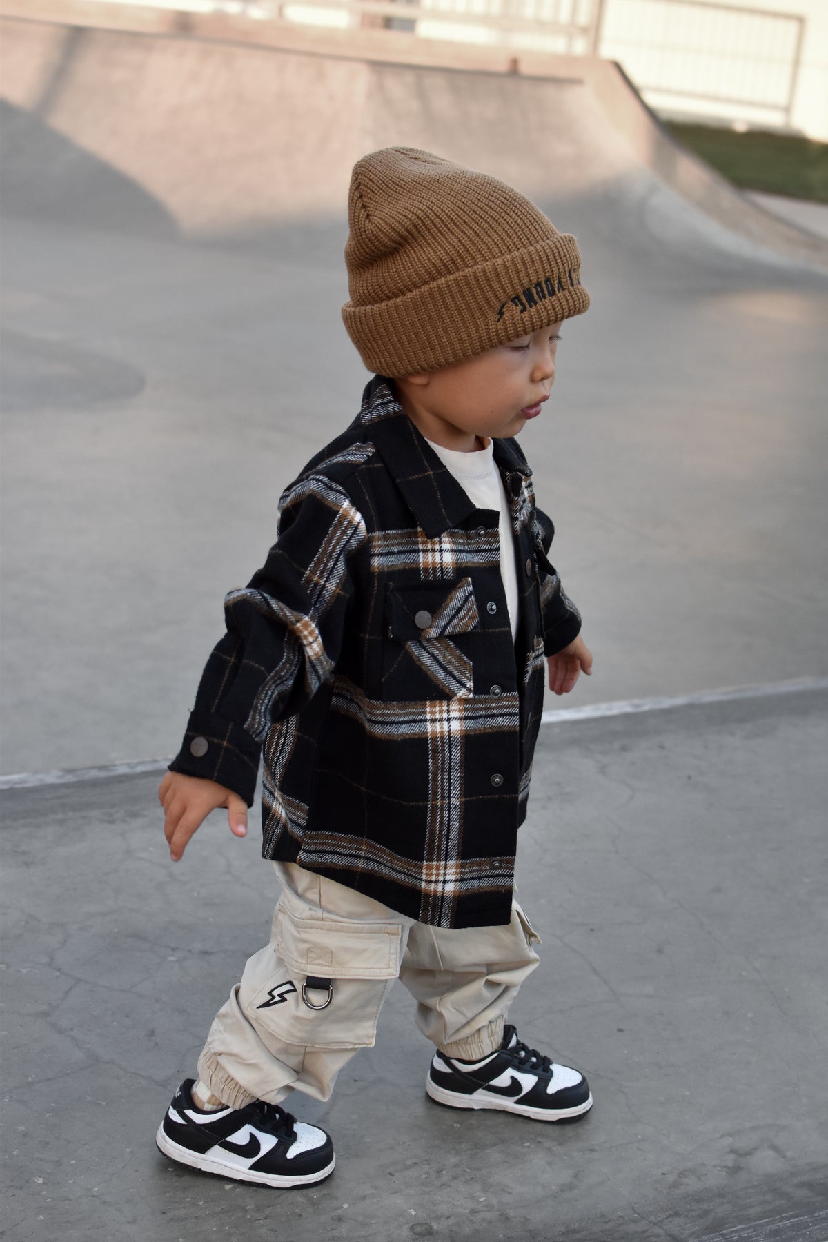 A toddler walking in a concrete skatepark wearing a brown knit beanie, a black and beige plaid button-up shirt, beige cargo pants, and black and white sneakers.