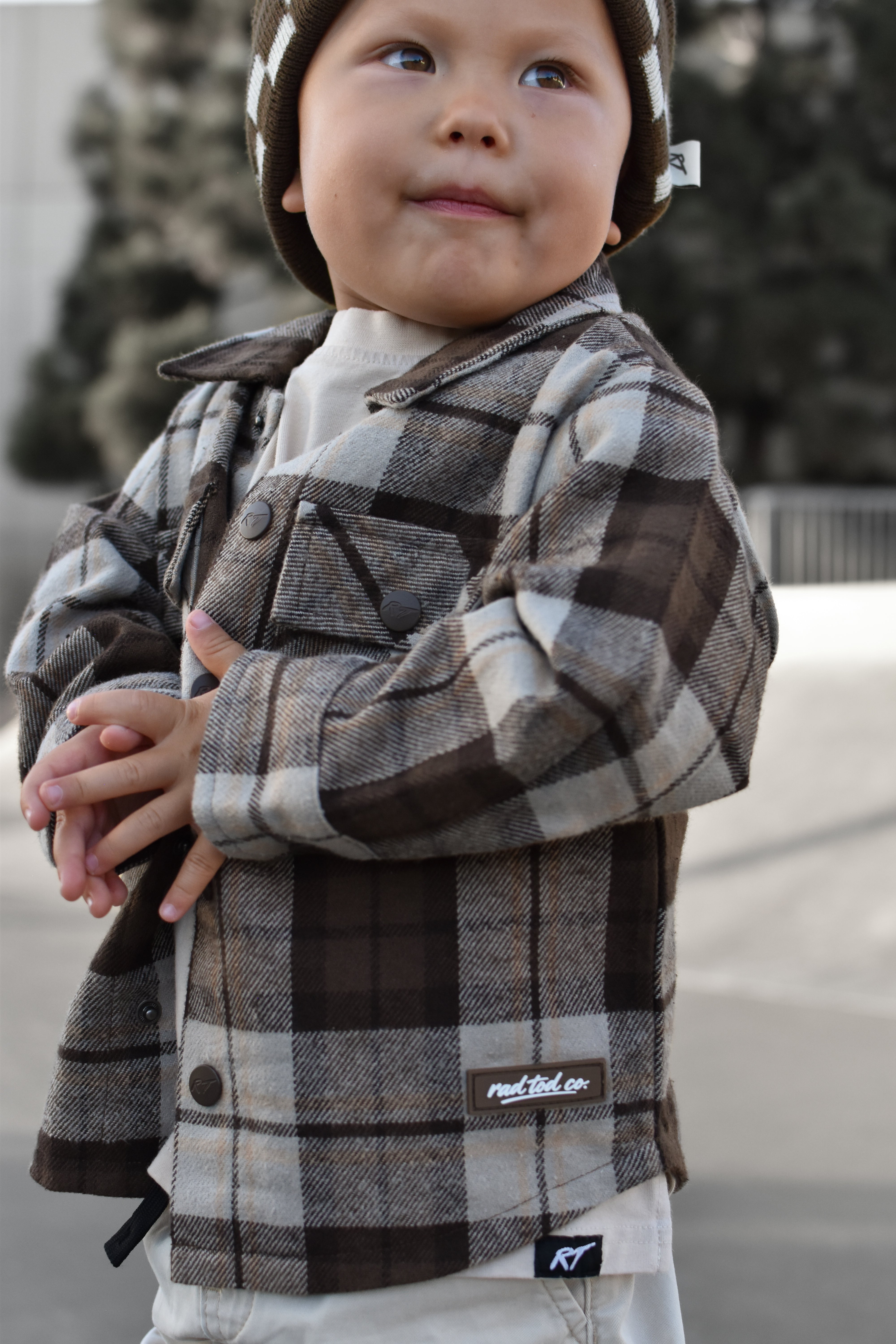 A toddler wearing a brown and beige plaid flannel button-up shirt jacket over a white t-shirt, paired with a matching plaid beanie. The jacket features a visible 'RAD TODDLER logo.