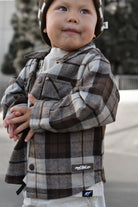 A toddler wearing a brown and beige plaid flannel button-up shirt jacket over a white t-shirt, paired with a matching plaid beanie. The jacket features a visible 'RAD TODDLER logo.
