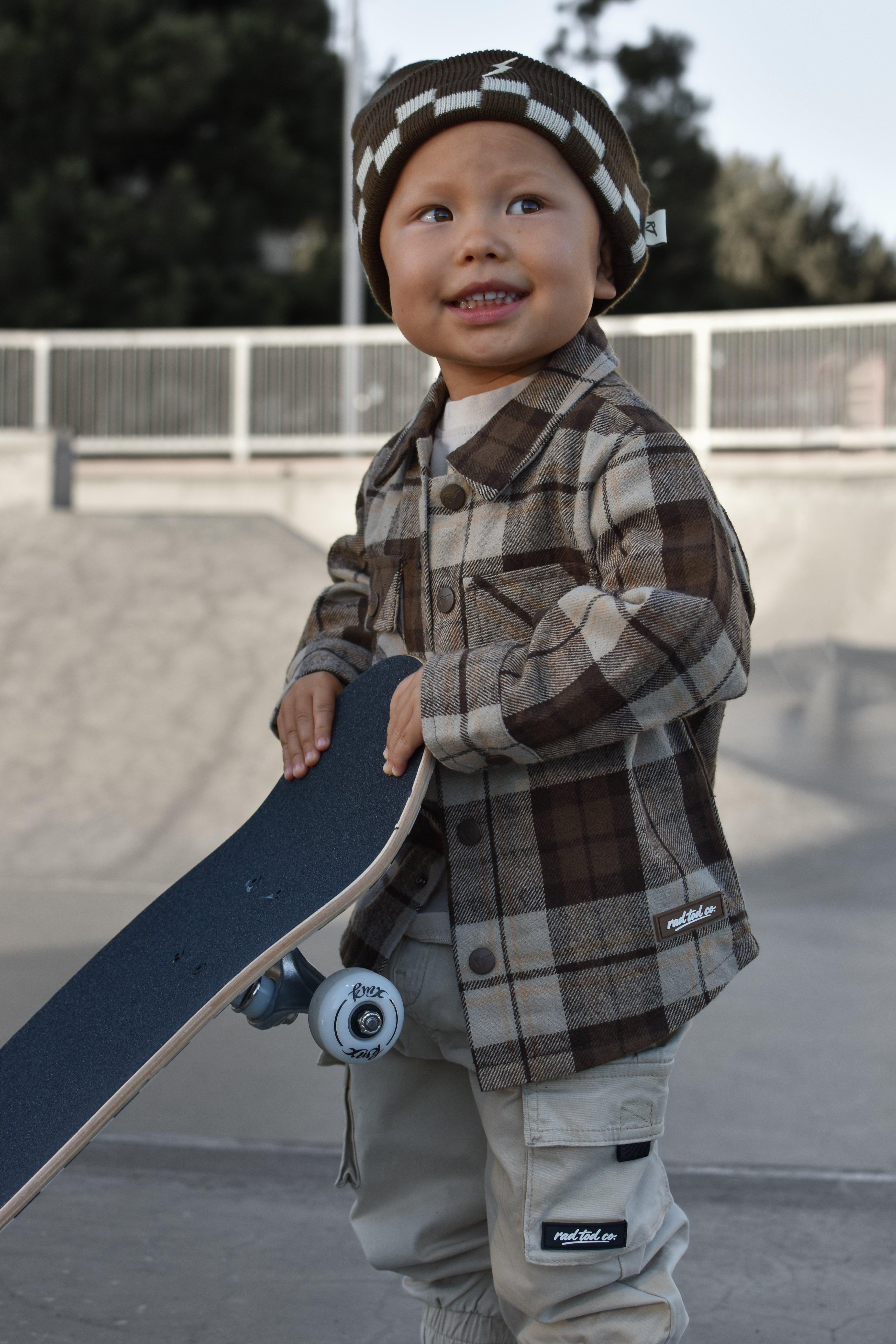 A young child wearing a brown and beige plaid button-up shirt, matching pants, and a checkered beanie hat, holding a skateboard at an outdoor skatepark.