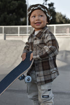 A young child wearing a brown and beige plaid button-up shirt, matching pants, and a checkered beanie hat, holding a skateboard at an outdoor skatepark.