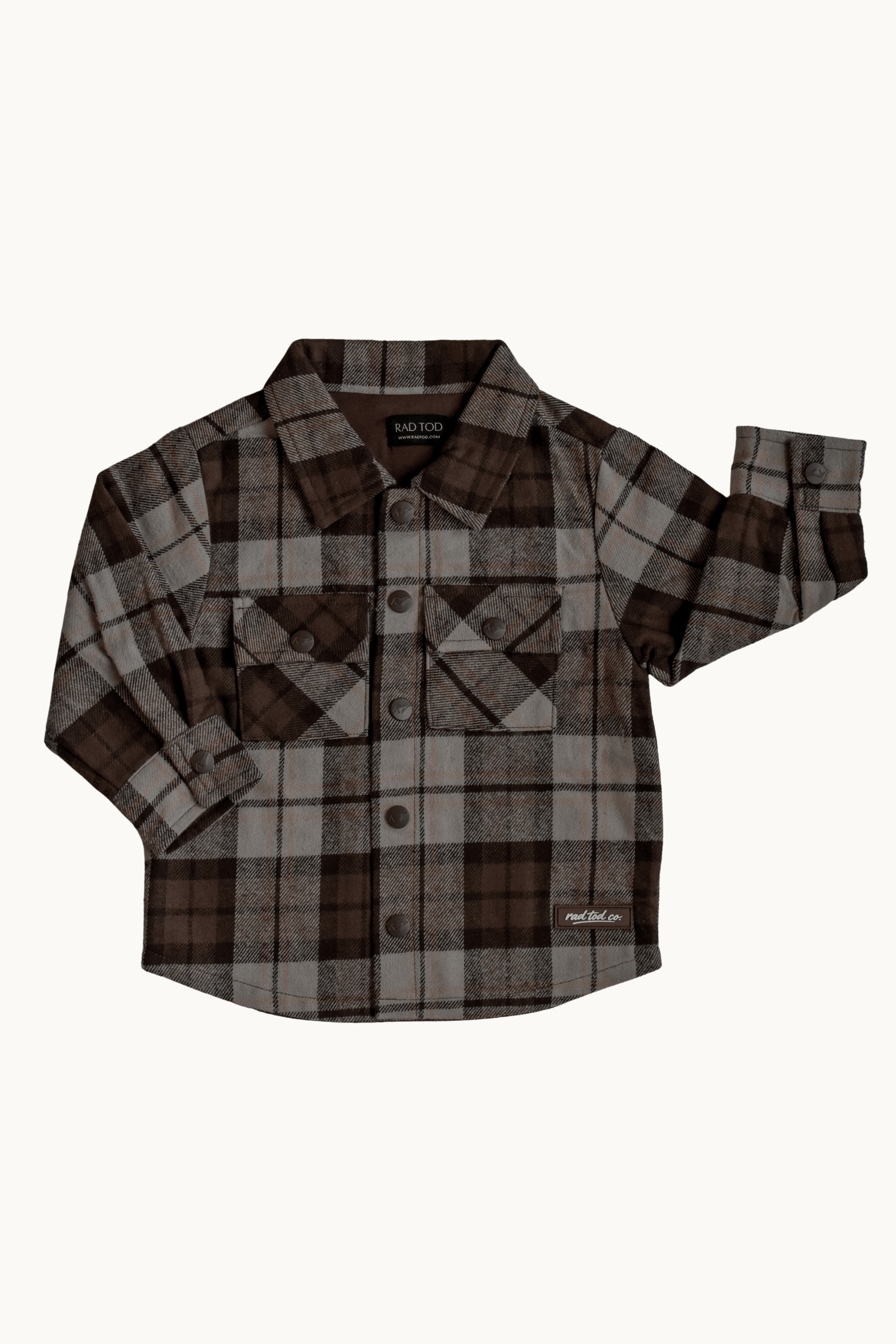 A long-sleeve plaid button-up shirt in brown, beige, and gray, featuring two chest pockets and a small white 'RAD TODDLER CO' logo on the lower right hem, displayed flat against a light off-white background.