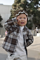 A toddler wearing a brown and beige plaid flannel shirt over a white t-shirt, paired with a brown and white checkered beanie, posing outdoors.